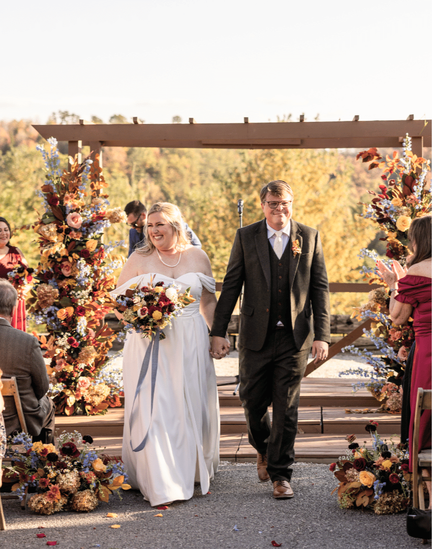 A bride and groom walking down the aisle at their wedding ceremony, holding hands and smiling, surrounded by colorful floral arrangements and guests applauding.