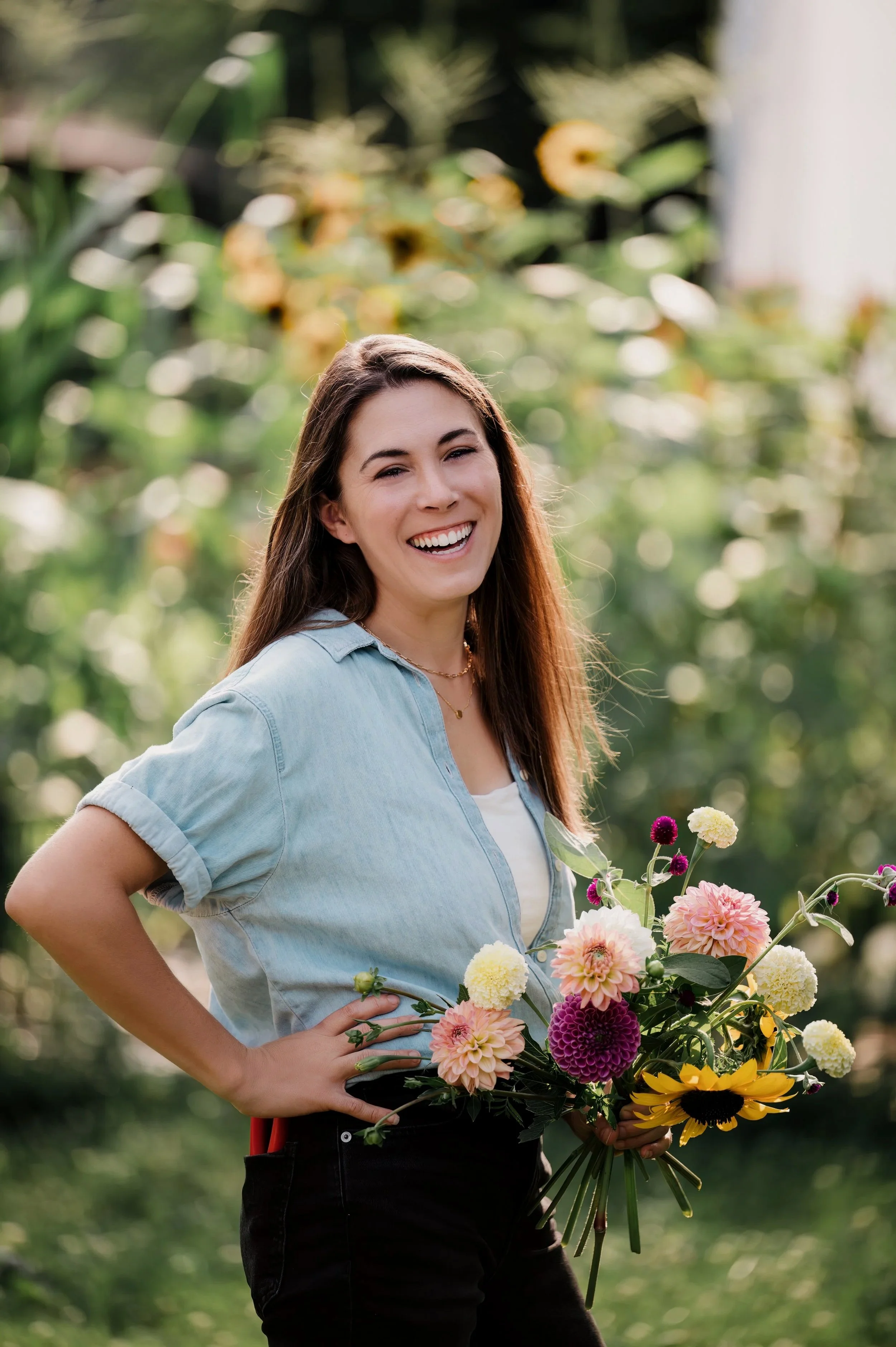 A young woman in a light denim shirt holding a colorful bouquet of flowers while smiling outdoors in a garden.