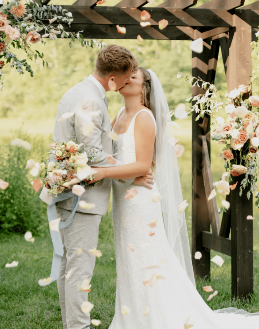 Couple in wedding attire sharing a kiss under an outdoor wooden arch decorated with flowers, with flower petals falling around them.