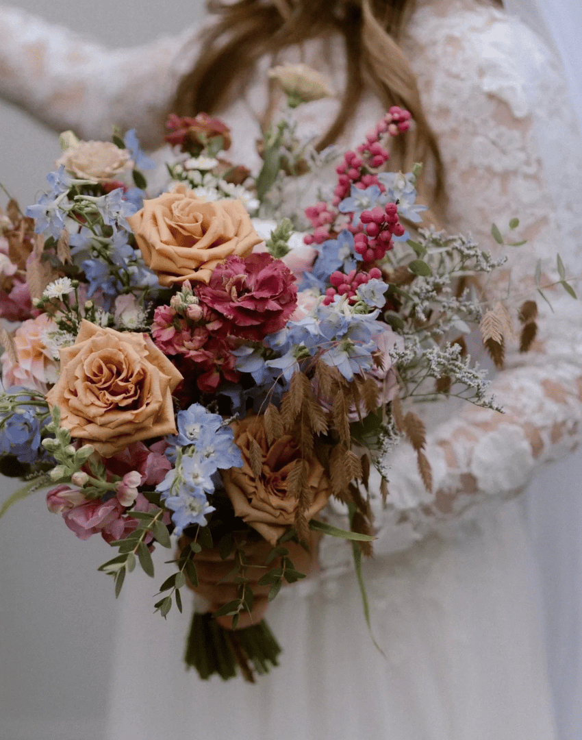 A woman in a lace dress holding a large bouquet of mixed flowers including beige roses, pink and red carnations, blue and purple delphiniums, and pink berries.