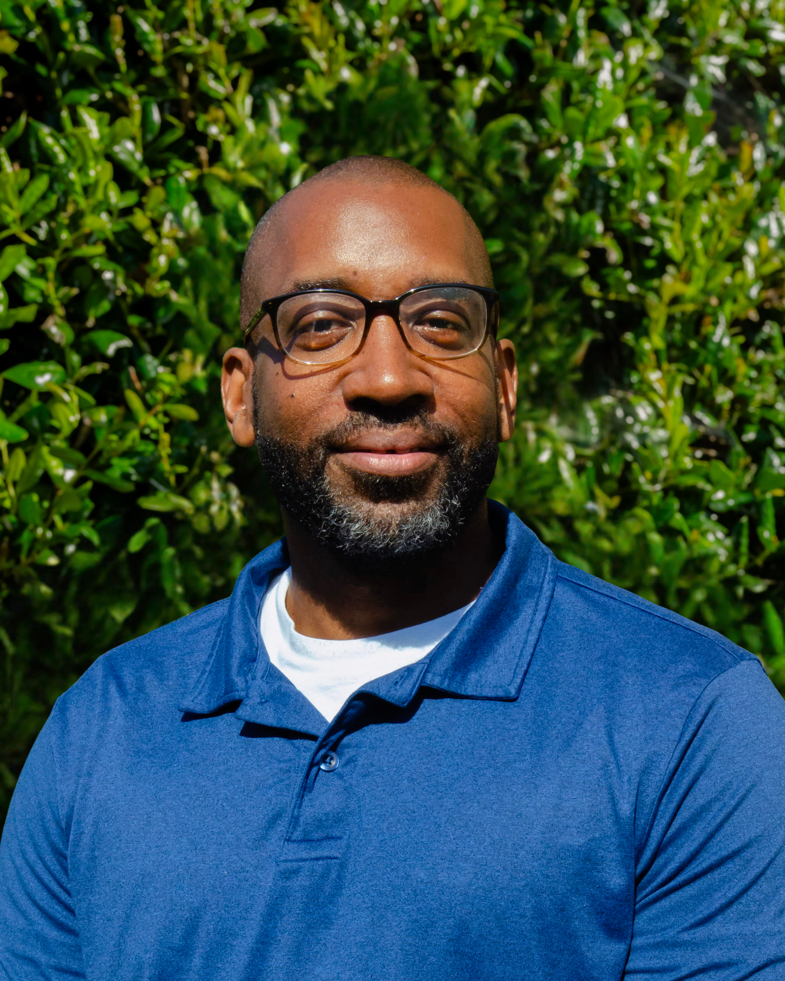 man outdoors, wearing a blue button-up shirt, with green foliage in the background.