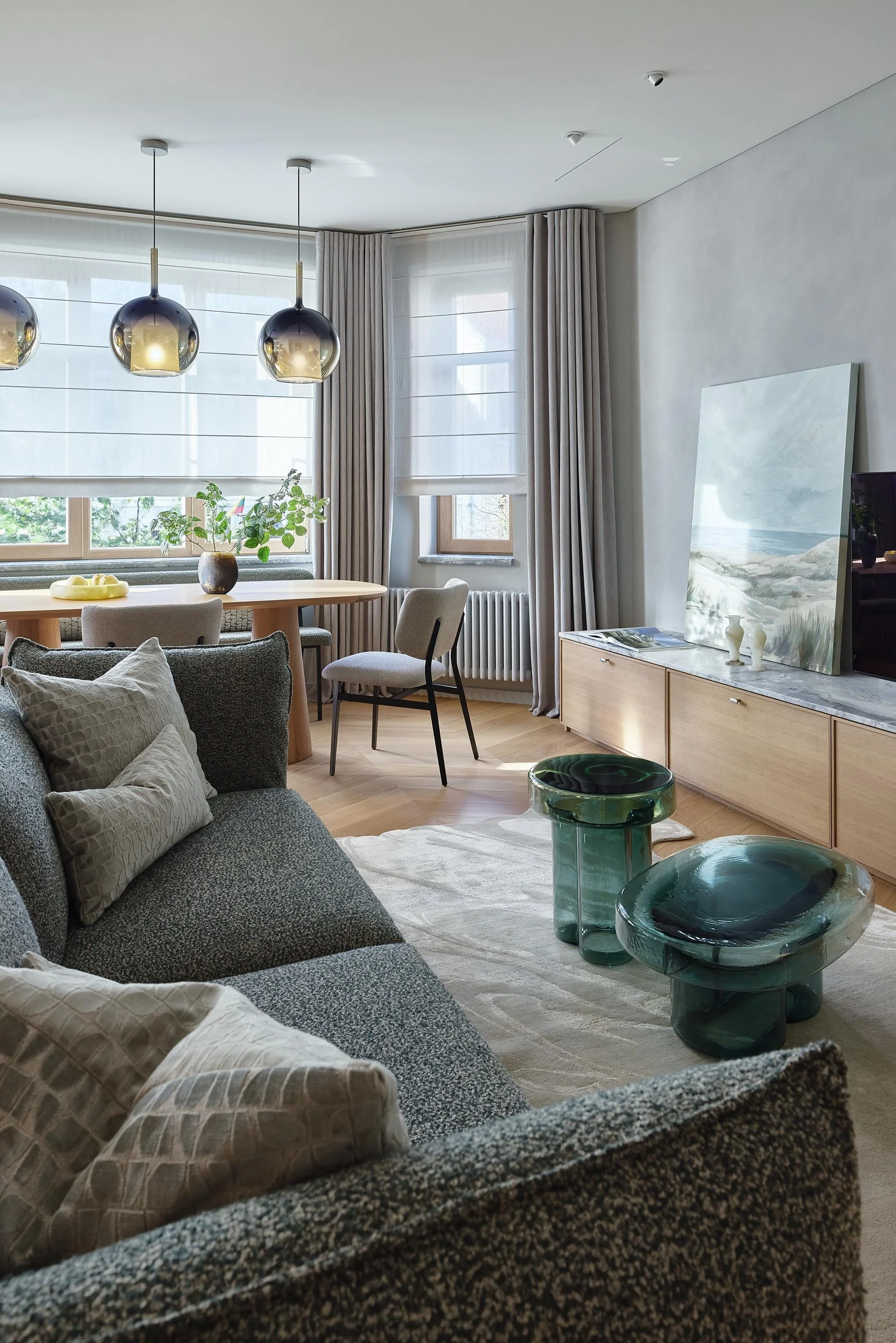 A living room with a gray sofa, beige pillows, a round wooden dining table, and modern glass tables. Large windows with white blinds and beige curtains. Wall art depicting a landscape, and contemporary lighting fixtures hanging from the ceiling.