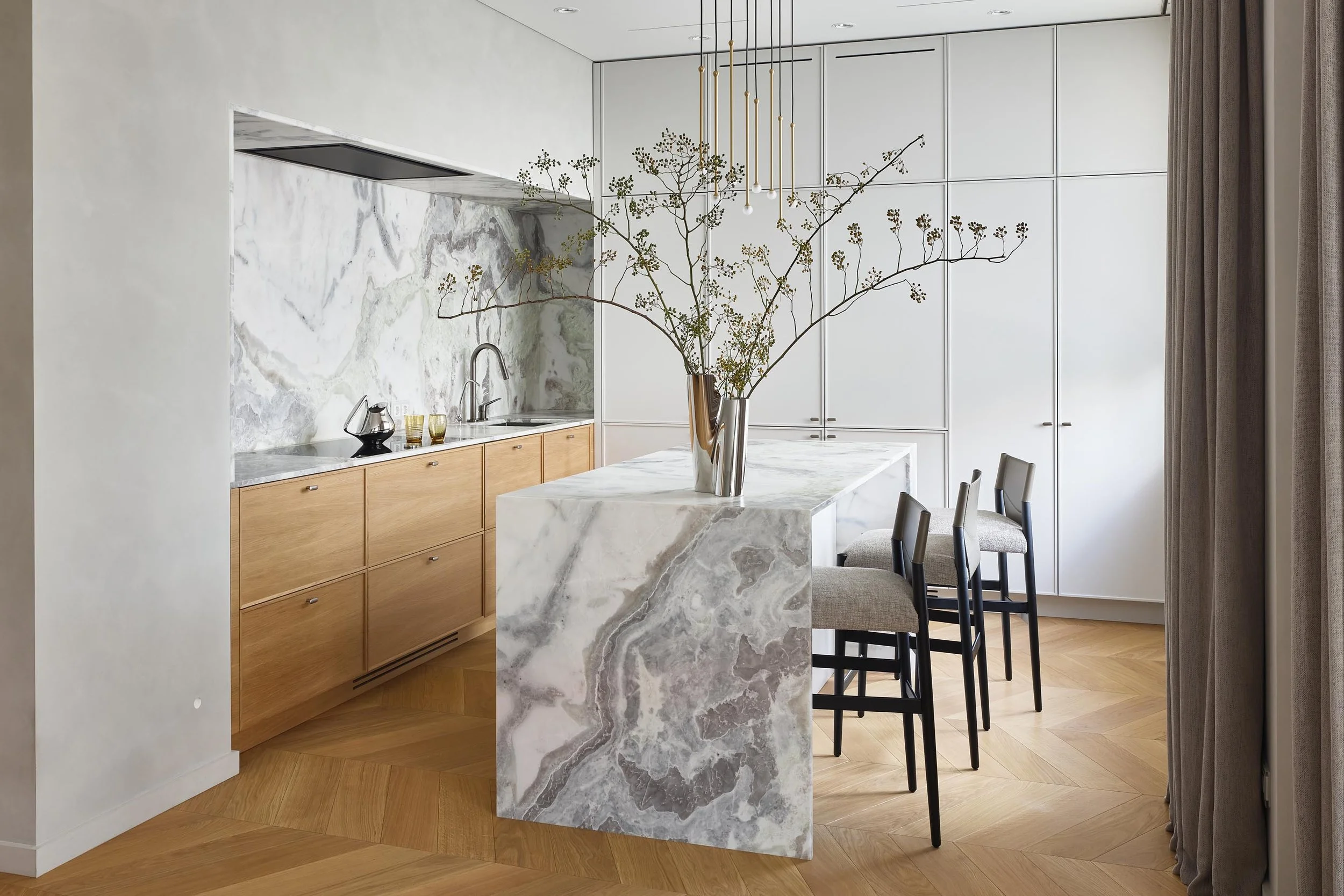 Modern kitchen with a marble kitchen island, light wooden cabinetry, white walls, and a large vase with a branch plant on the island. There are black chairs around the island and white ceiling lights with gold accents.
