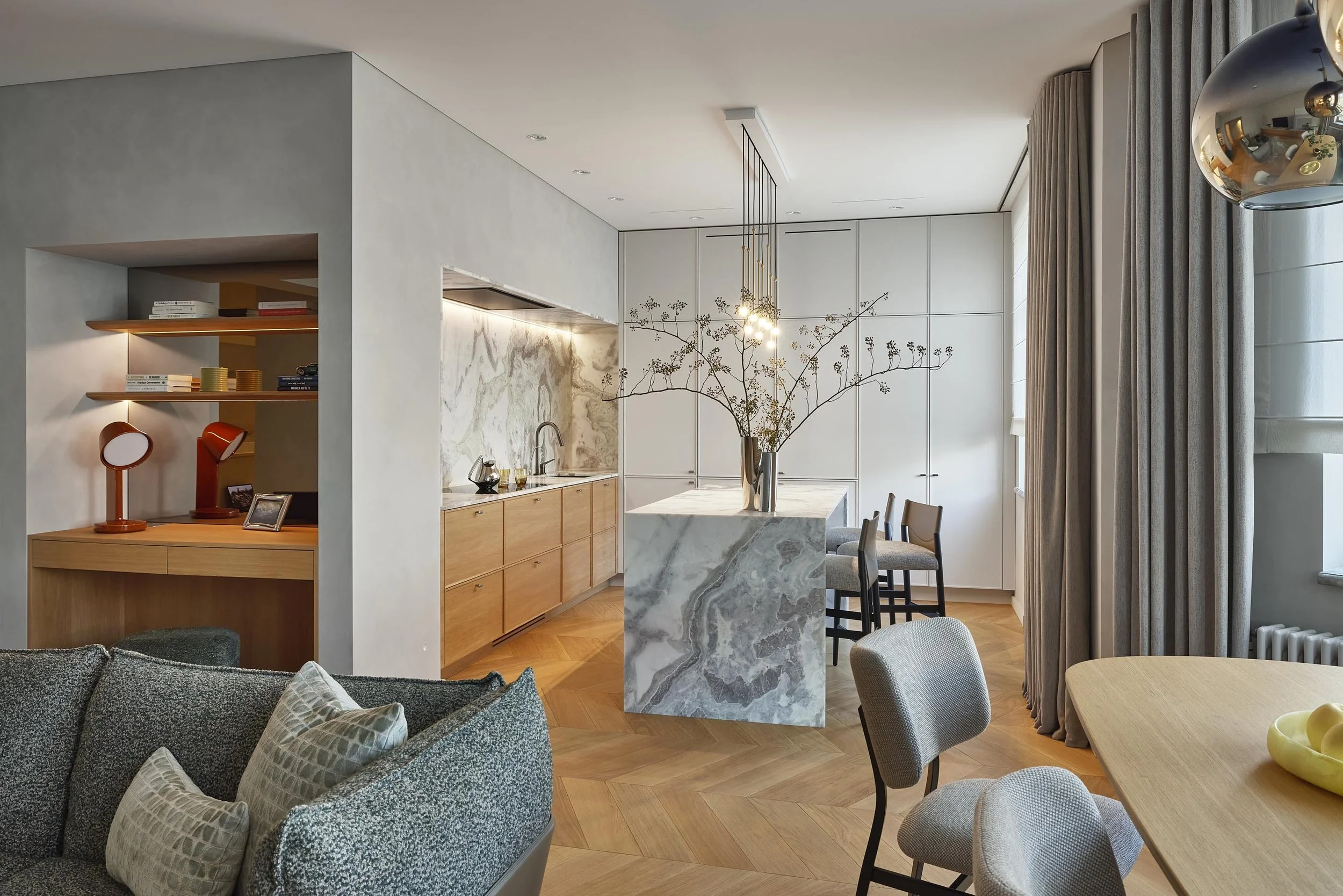 A marble waterfall island stands at the centre of the kitchen, while the veined stone splashback glows softly under the shelf lighting. Light oak cabinetry, chevron parquet and a single branch in a silver vase keep the whole room quiet.