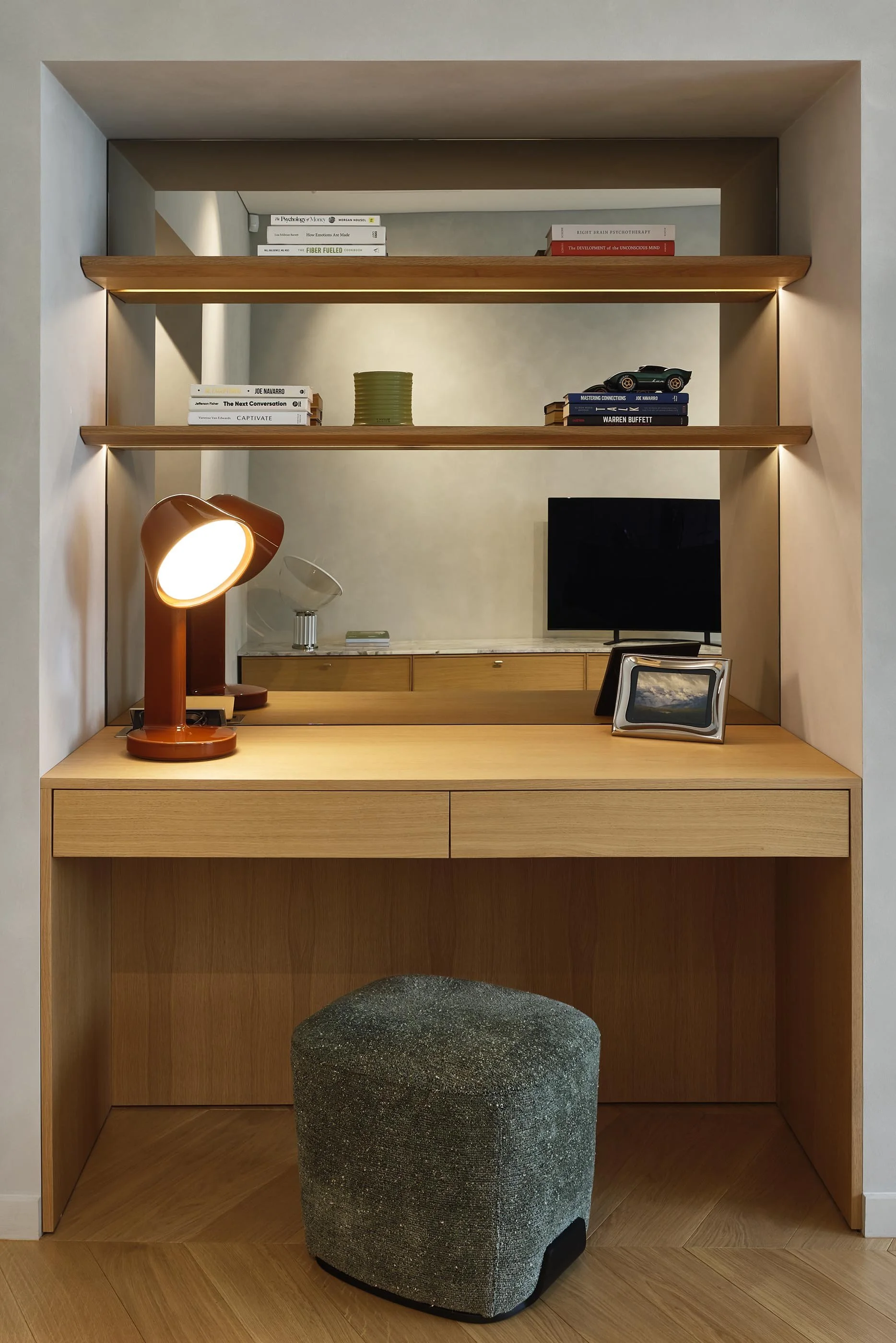 A built-in oak desk with two slim drawers sits in a plastered niche, lit from above by floating shelves with concealed lighting. The mirrored back panel doubles the light and reflects the living room, while a rust-red ceramic lamp adds the single not