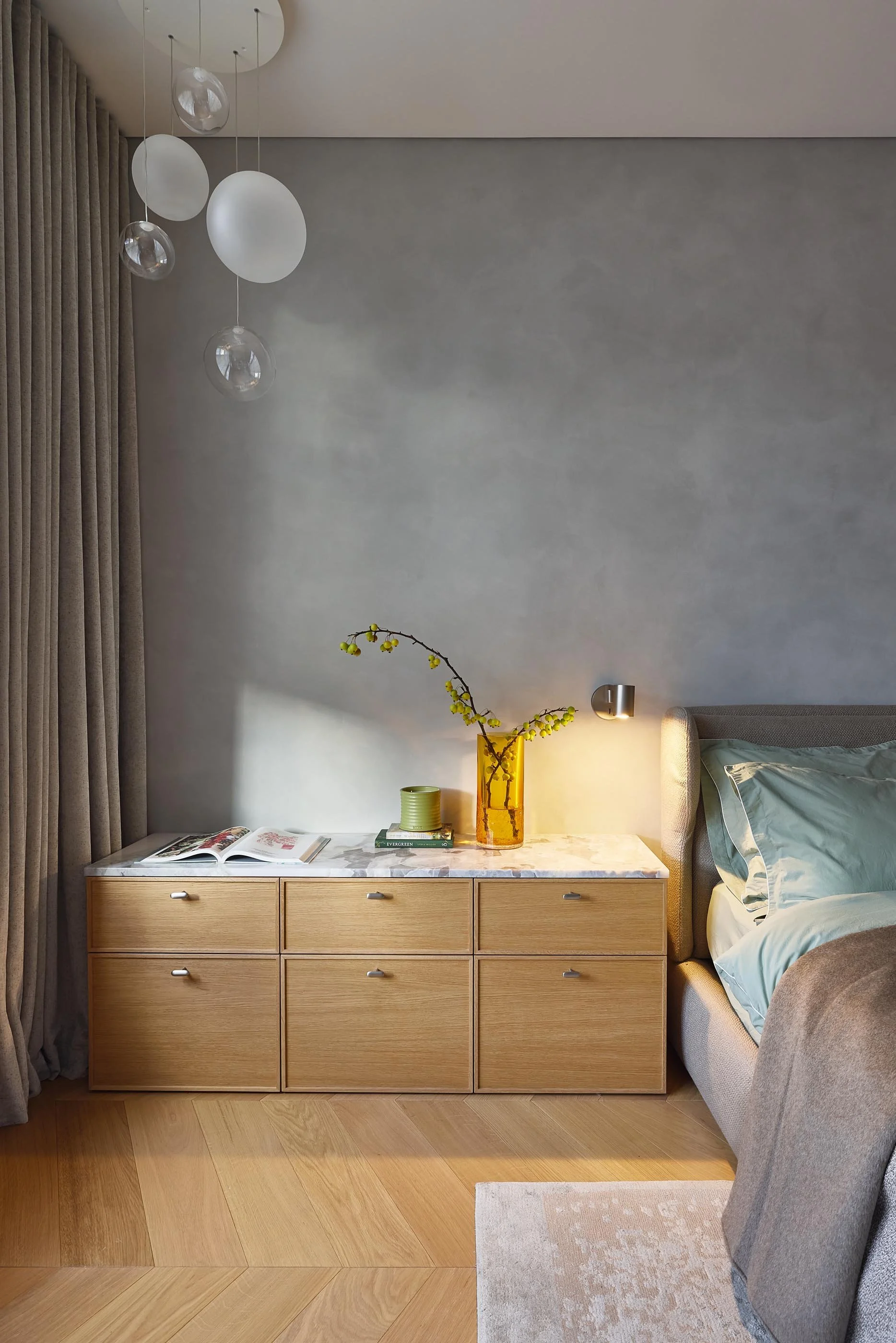 Bedroom with a bedside table, a bed, and a wall-mounted light. The bedside table has a yellow vase with branches and yellow berries, a green ceramic cup, and an open magazine. There are beige curtains and wood flooring.