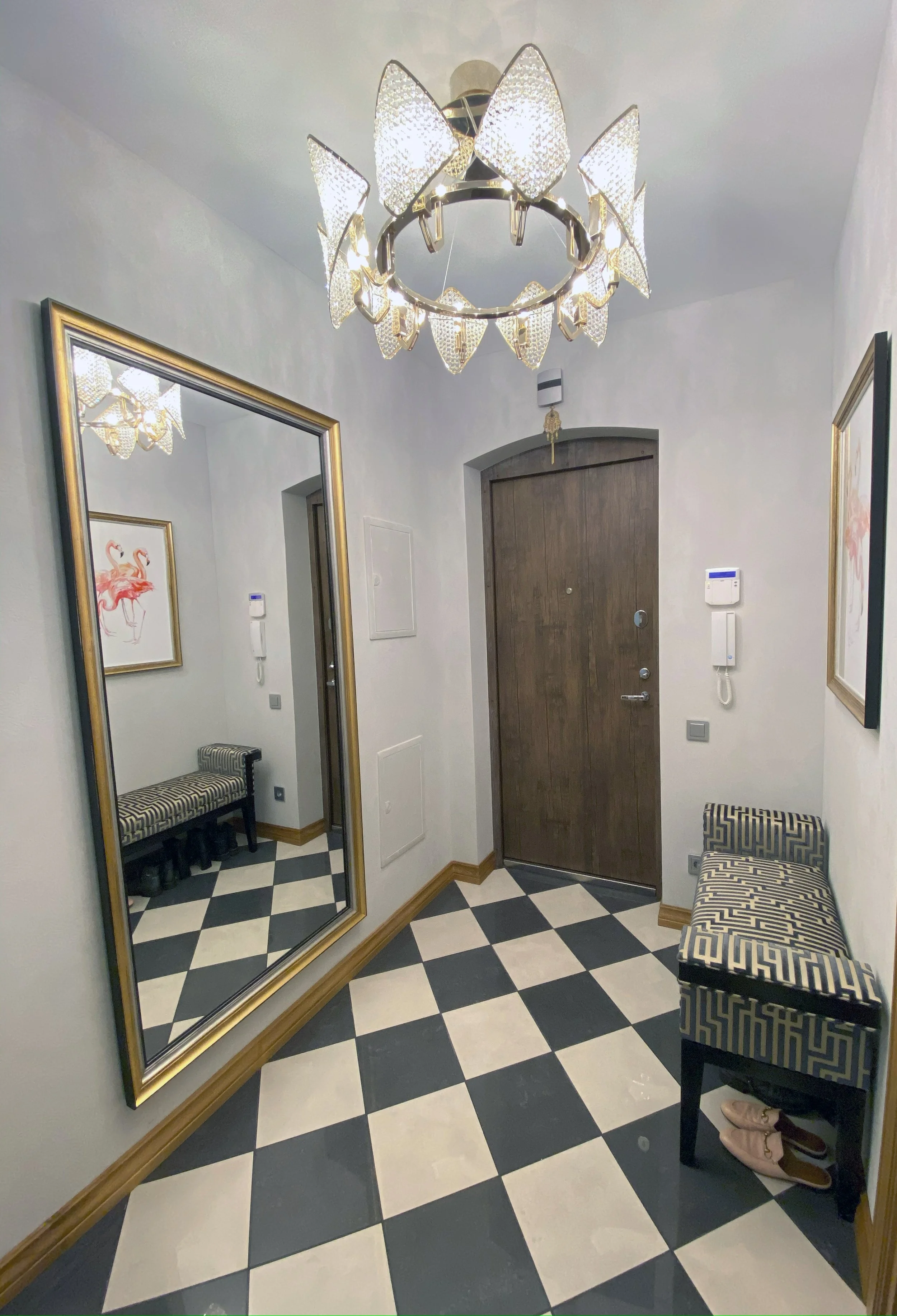 A hallway with a black and white checkered floor, a large mirror with a gold frame, seating, a wooden door, and decorative wall art of pink flamingos.