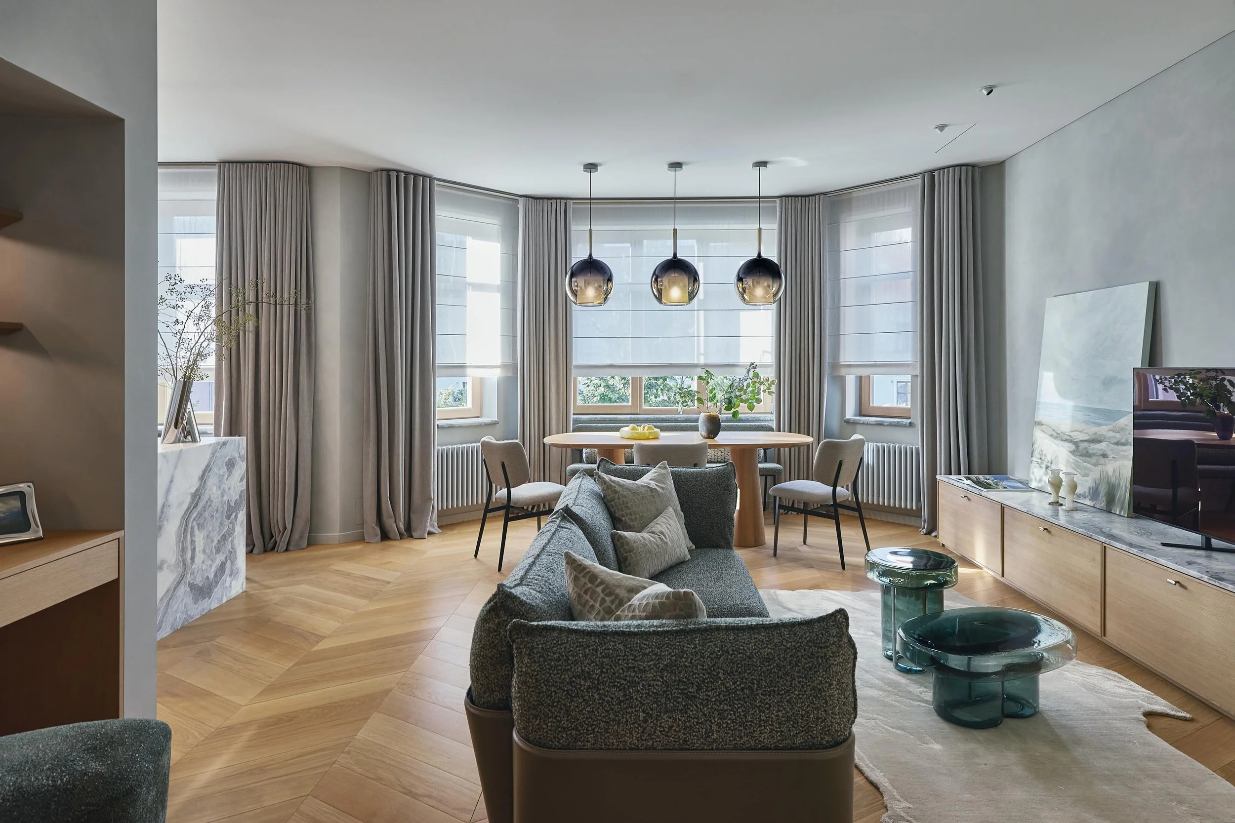 Modern living room with large windows, beige curtains, a round wooden dining table with six chairs, gray sofa with pillows, abstract painting, TV on a wooden stand, and glass coffee tables.