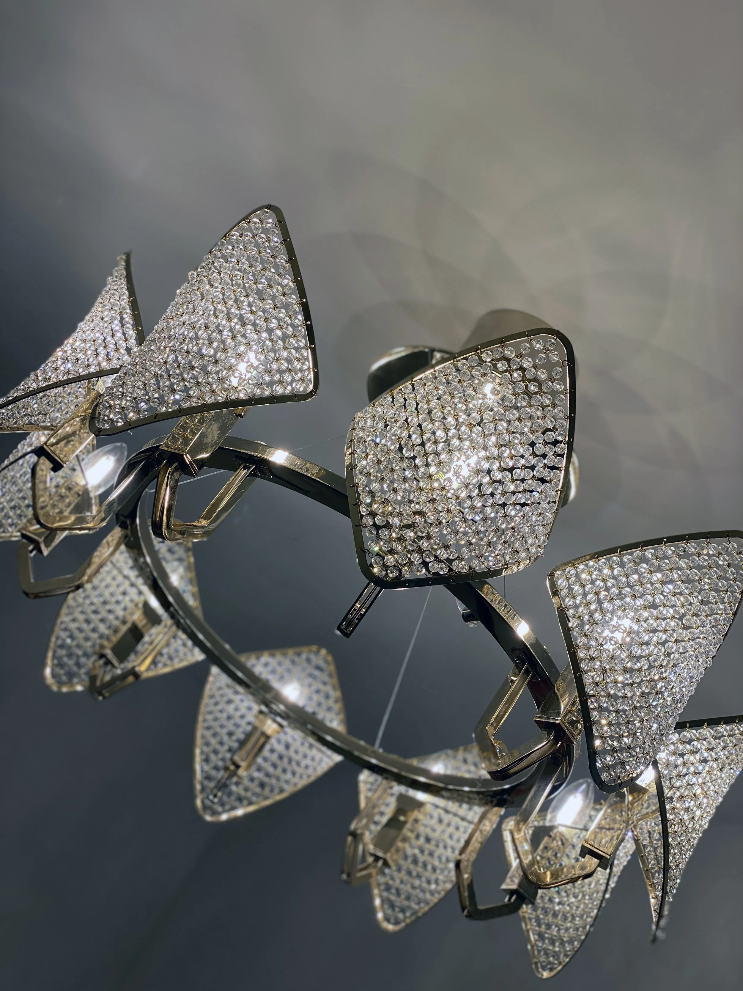 A close-up view of a decorative butterfly-shaped chandelier with numerous small crystals, illuminated and hanging against a dark gray background.