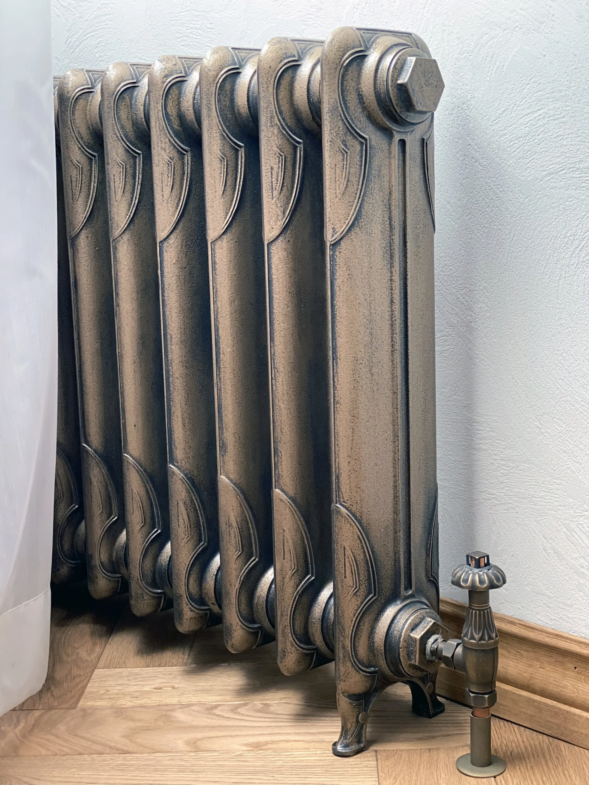 An antique-style cast iron radiator with a gold and black finish, positioned next to a small decorative lamp on a wooden floor with a white textured wall.