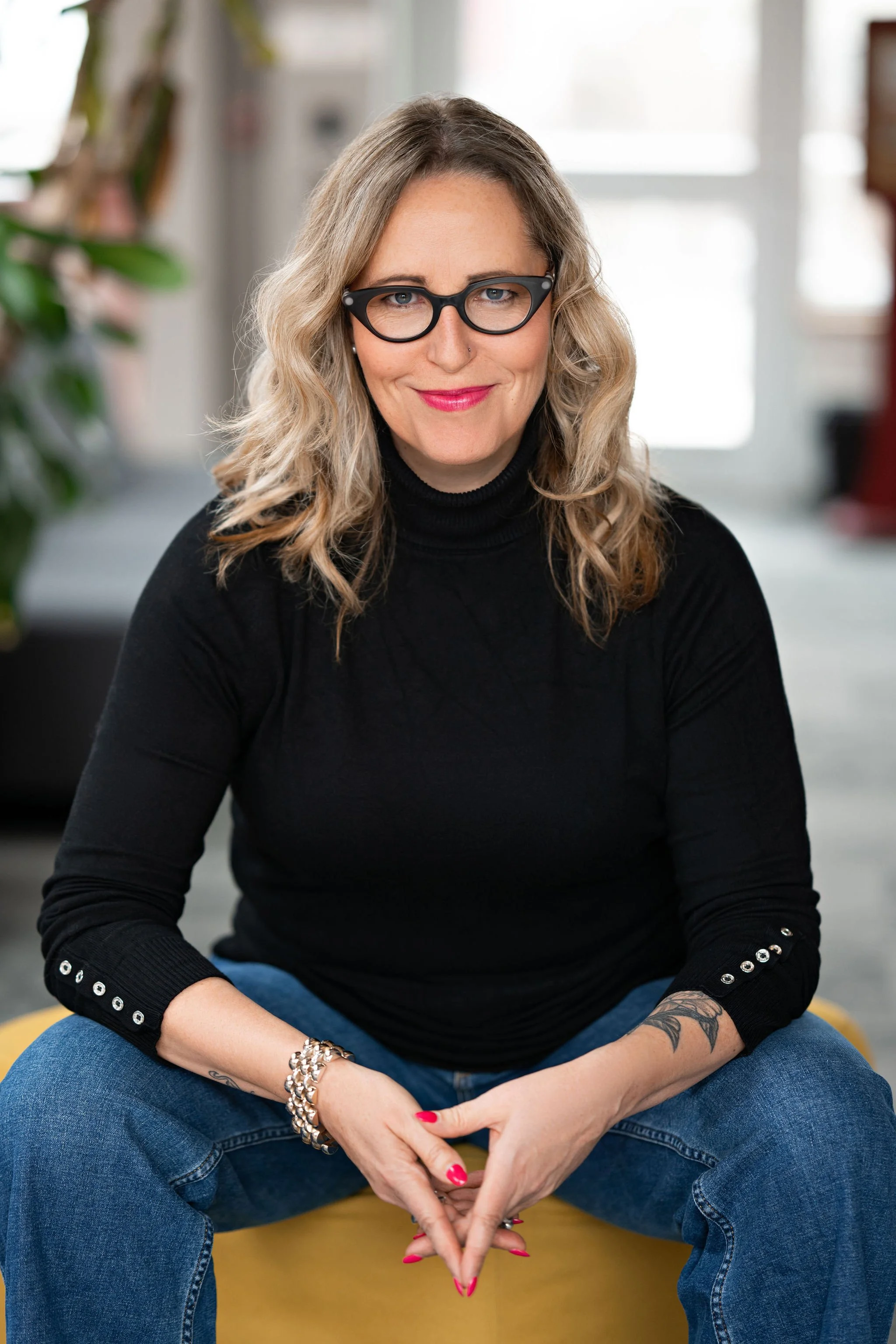 A woman with wavy blonde hair, wearing black glasses, a black turtleneck with button details on the sleeves, and blue jeans, sitting on a yellow chair in a bright, modern indoor setting.