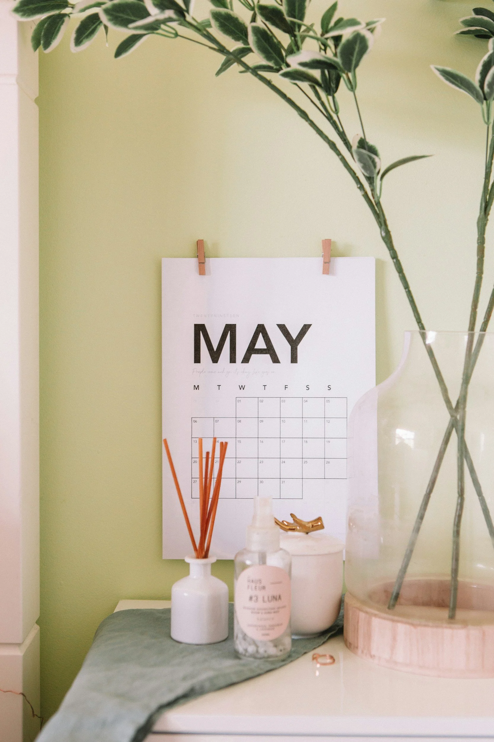 A May calendar hung on a light green wall, with a potted plant in a glass vase and decorative items on a table below.