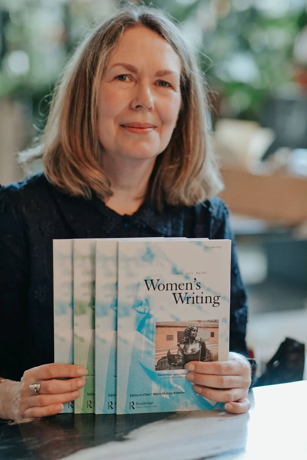 A photo of a seated woman (Louise Duckling) against a blurred green background, wearing a dark blue blouse, and holding four issues of the journal Women's Writing.
