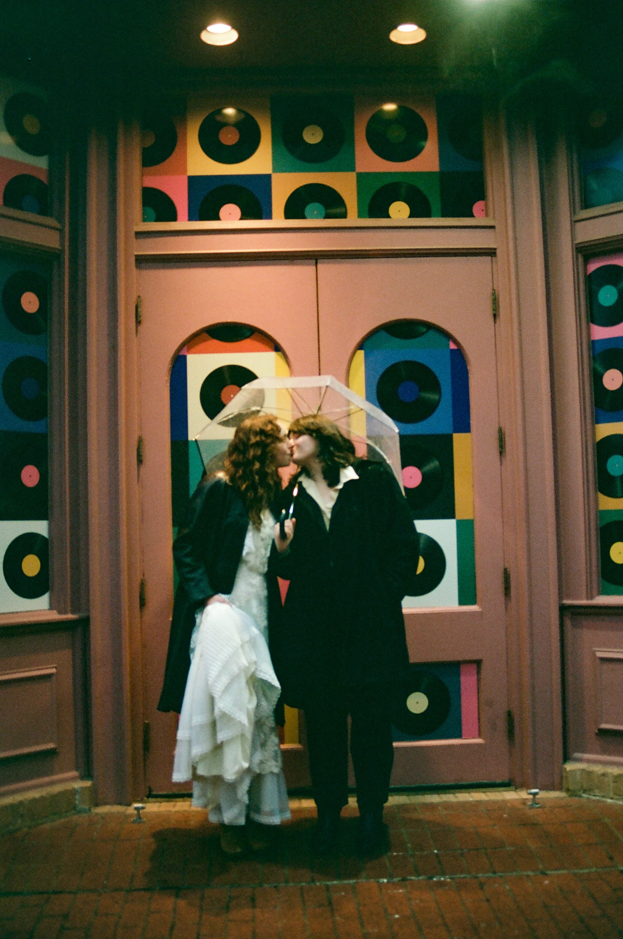 Two women with curly hair sharing a kiss under an umbrella outside a colorful building decorated with vinyl record patterns.