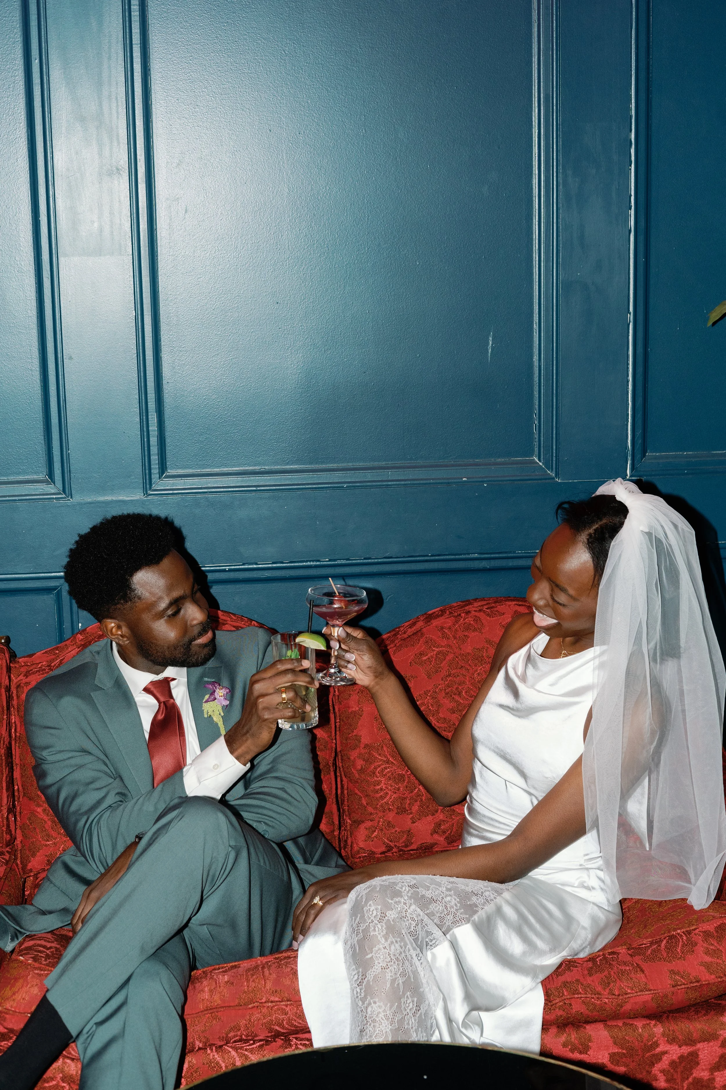 A couple, dressed in wedding attire, sitting on a red couch, clinking cocktails and smiling at each other in a room with blue walls.