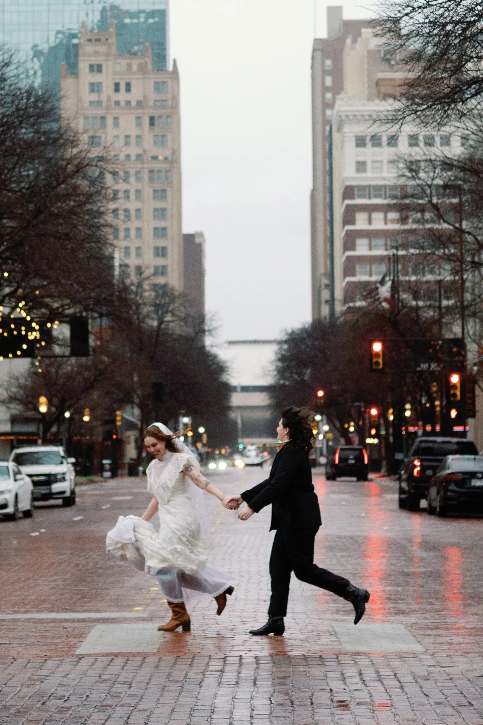 Two women holding hands crossing a city street at dusk, one in a white wedding dress and boots, the other in a black suit and boots, with tall buildings and traffic lights in the background.