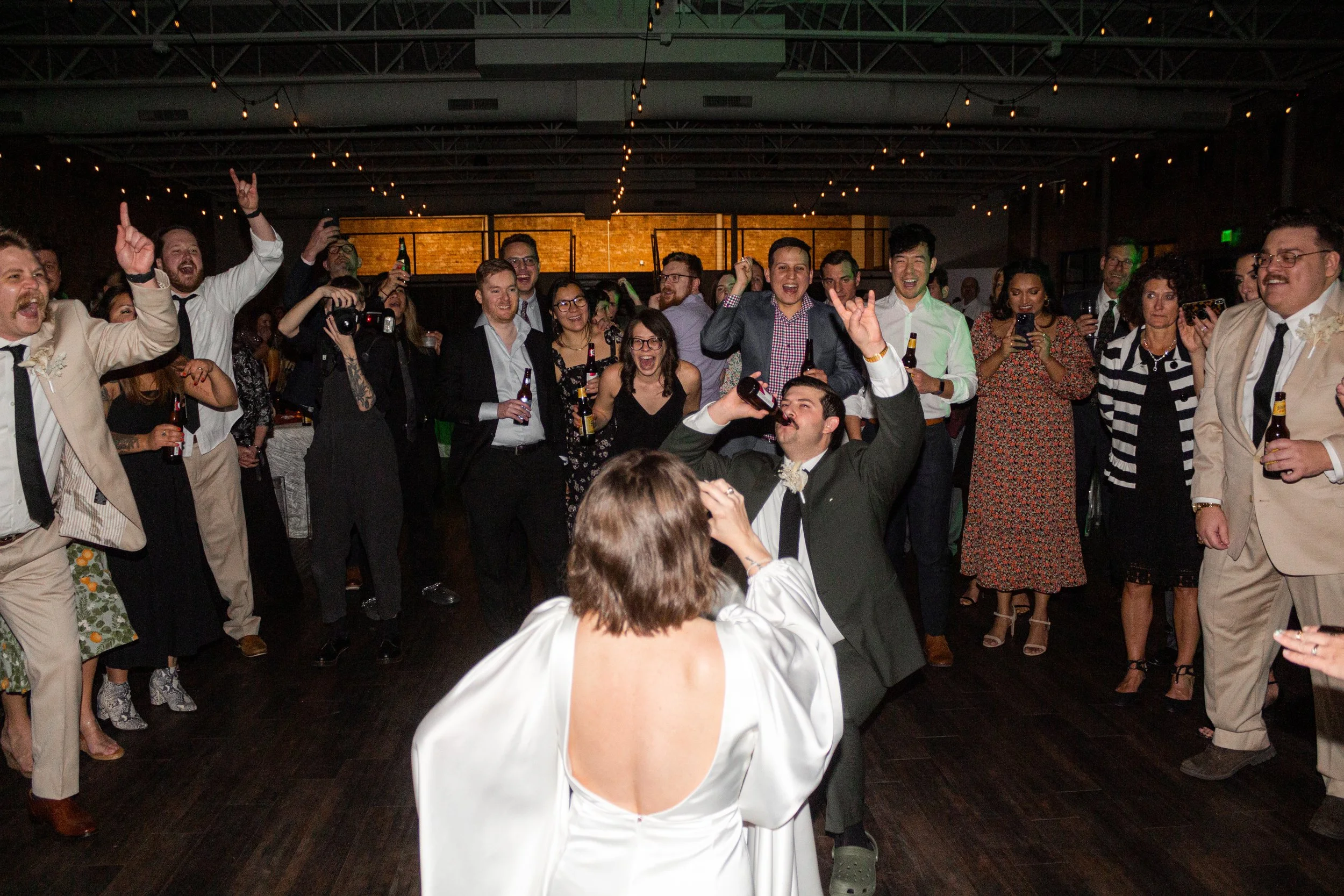 A wedding reception with newlyweds dancing in front of guests who are smiling, taking photos, and raising their drinks.