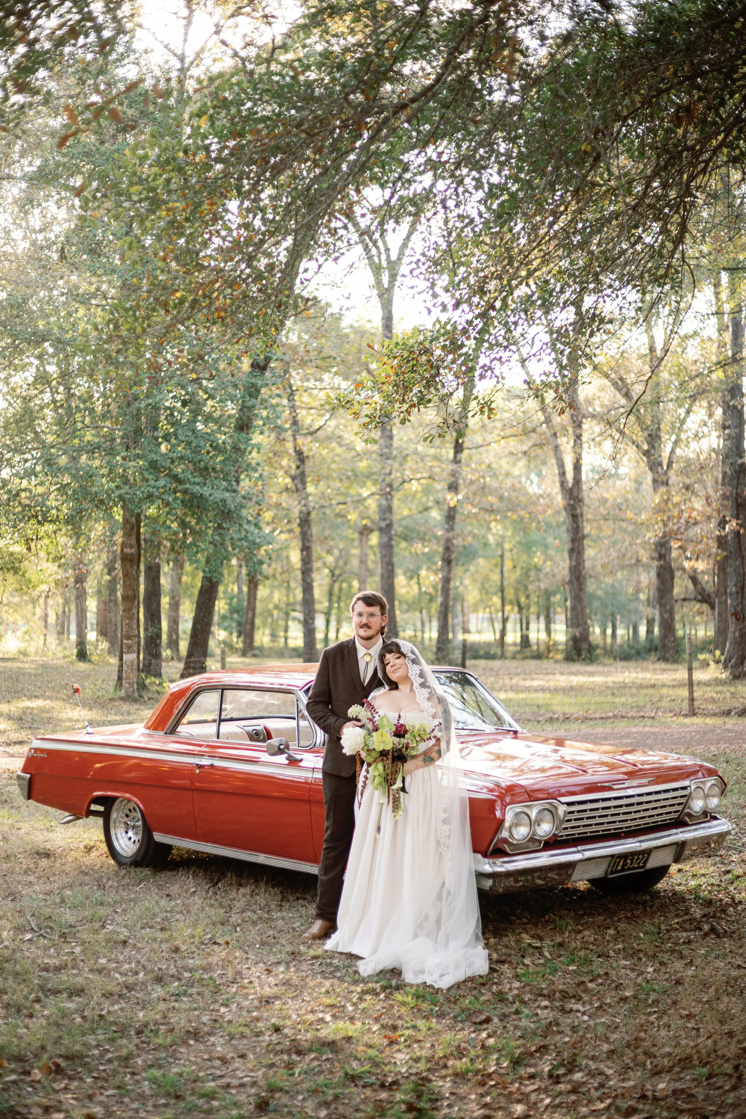 Newlyweds pose together for a picture infant of a vintage car during their outdoor wedding reception.