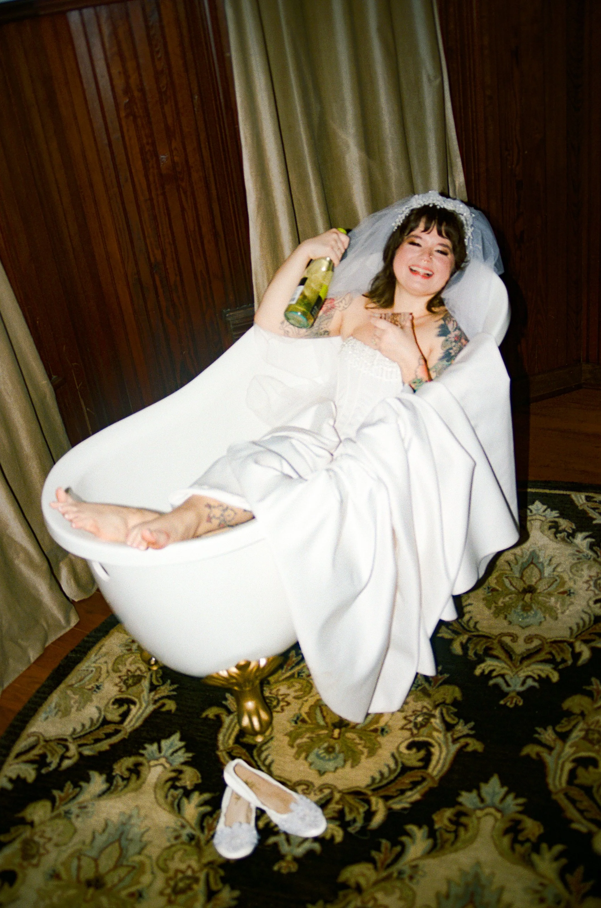 A woman in a wedding dress lying in a bathtub smiling and holding a bottle of champagne.
