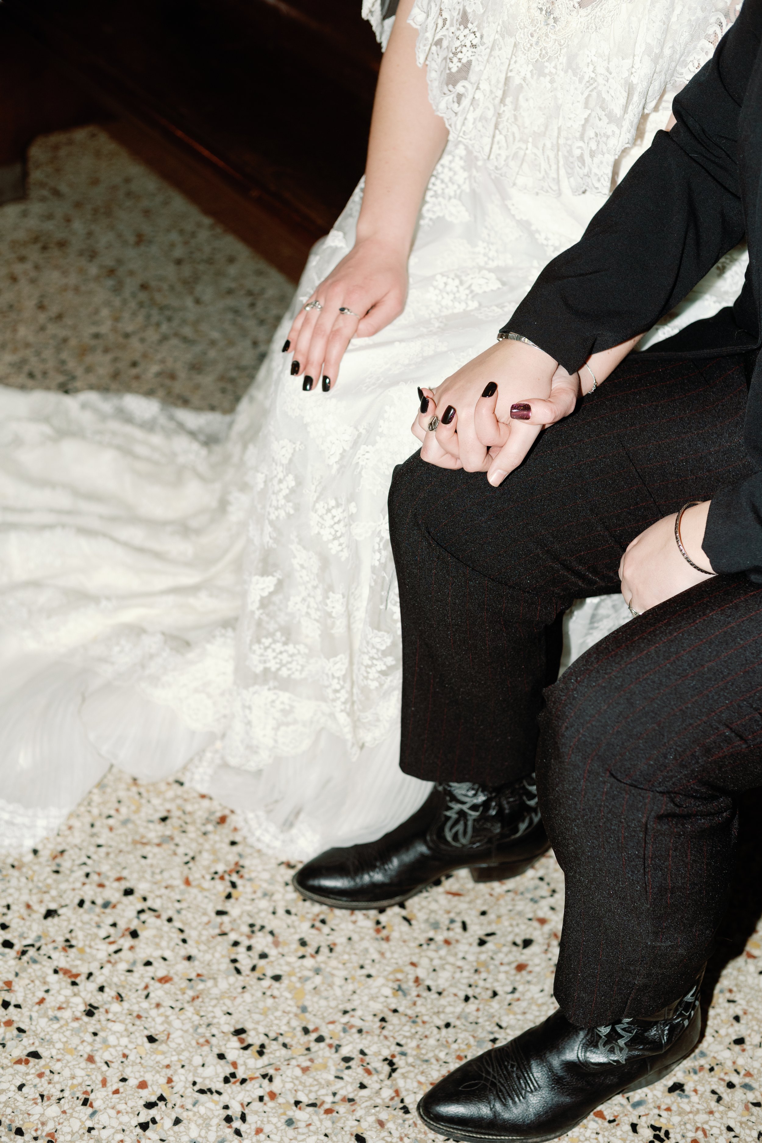 Close-up of Newlyweds' hands, dressed in wedding attire, sitting on a speckled floor.