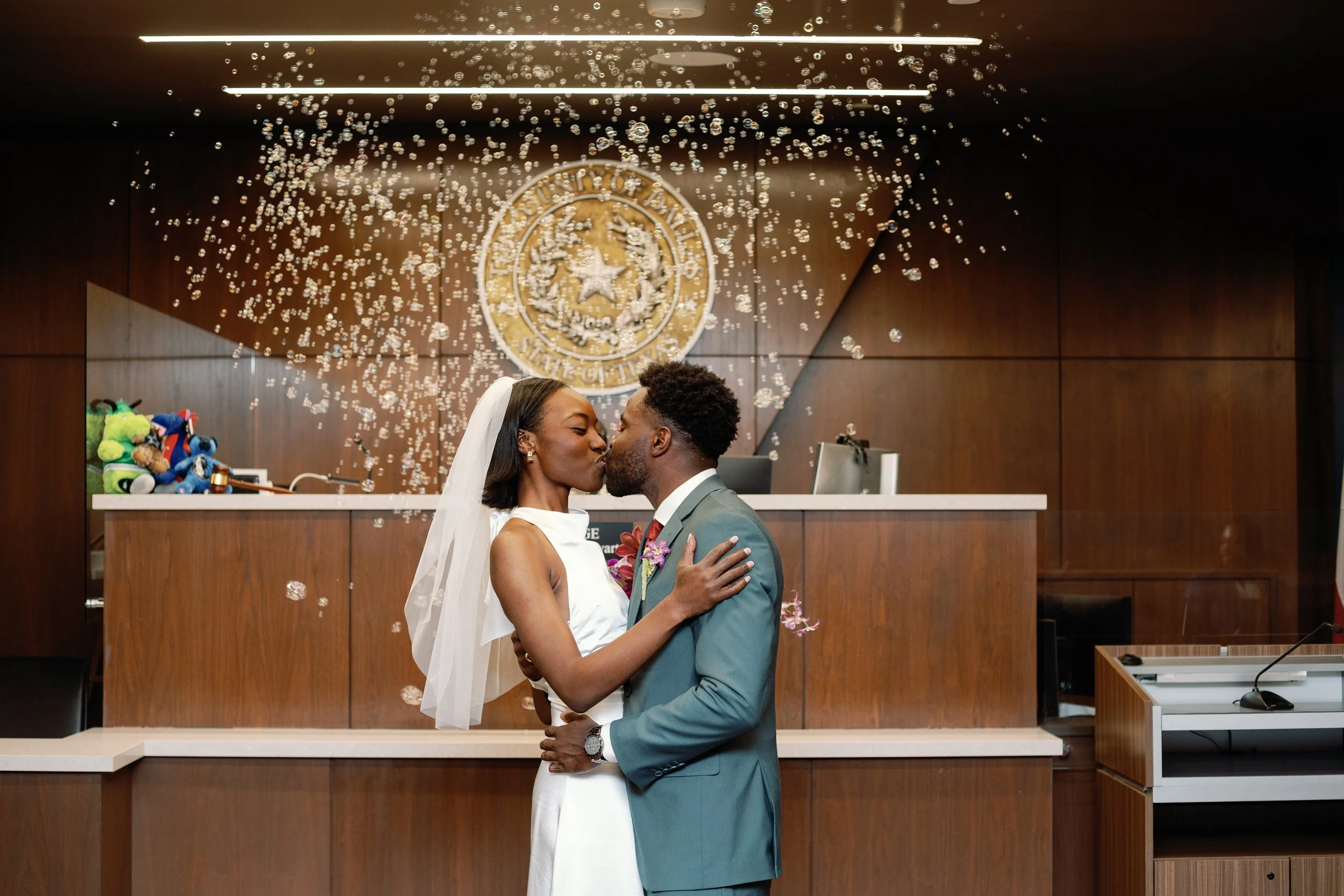 Newlyweds kiss during their wedding ceremony, with bubbles floating around them inside a courtroom.