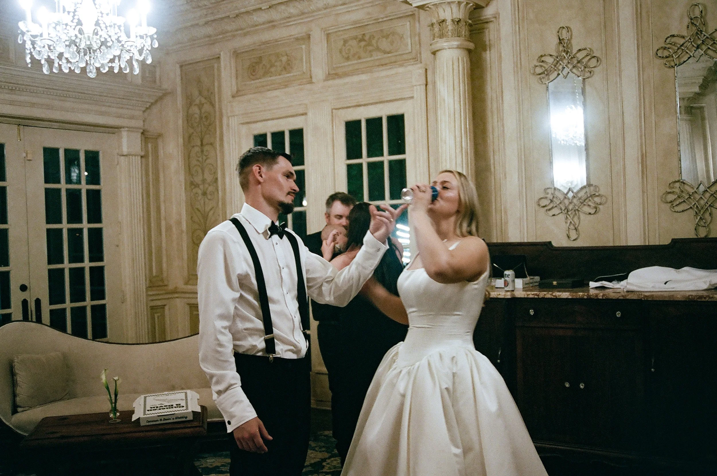 Newlyweds drink together inside their wedding venue during the reception on their wedding night. One of them is holding a beer while the other chugs it.
