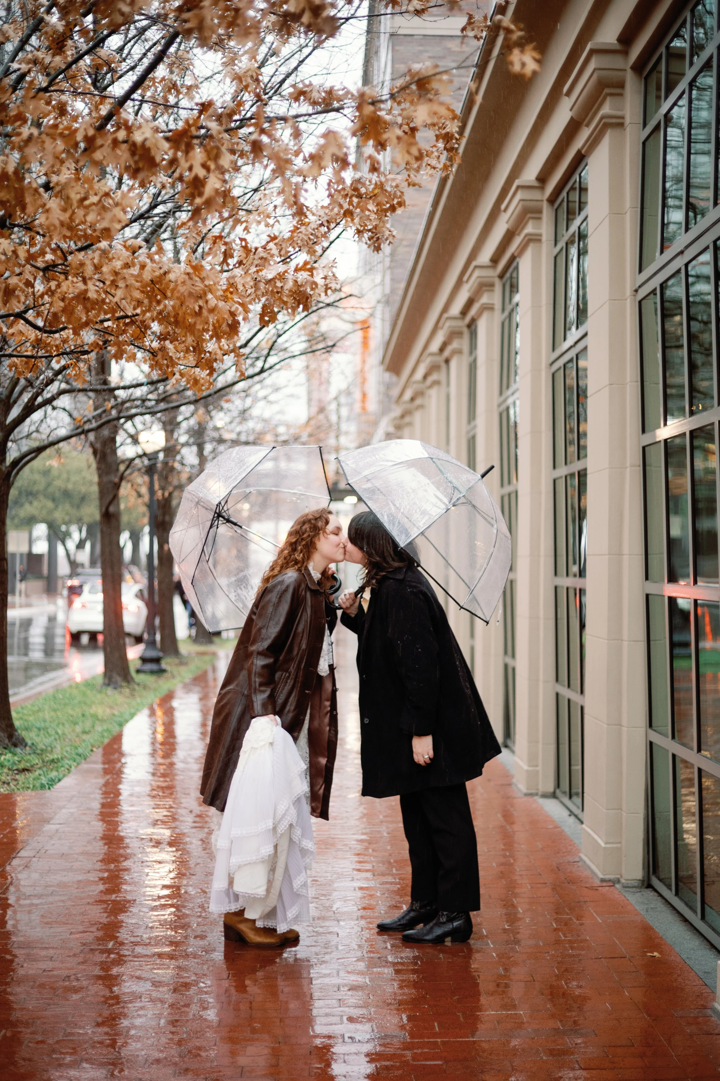 Two women sharing a kiss under umbrellas on a rainy day on a city sidewalk with autumn trees.