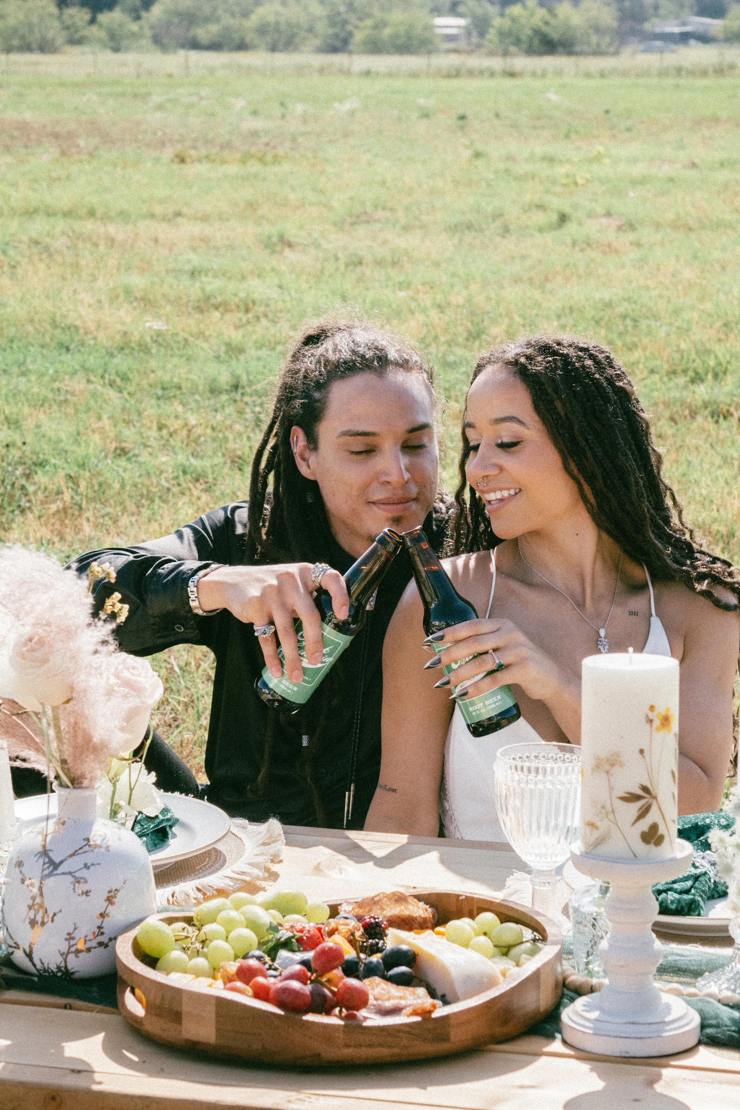 Newlyweds celebrating outdoors at a table with fruit, flowers, a candle, and drinks, clinking beer bottles together.