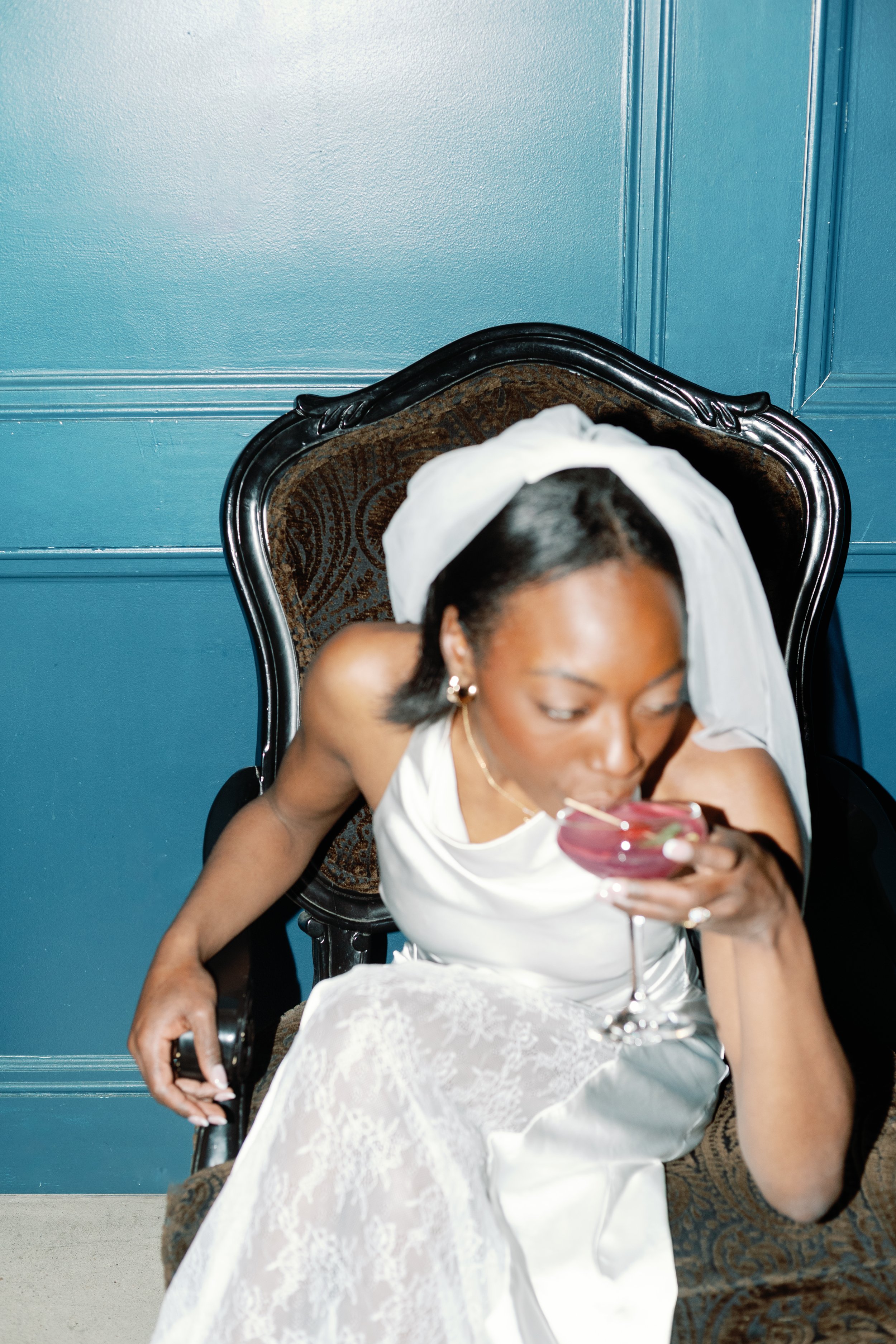 A woman wearing a wedding gown and veil is sitting on an ornate dark wood chair with patterned upholstery, drinking a pink cocktail in a coupe glass, against a blue paneled wall.