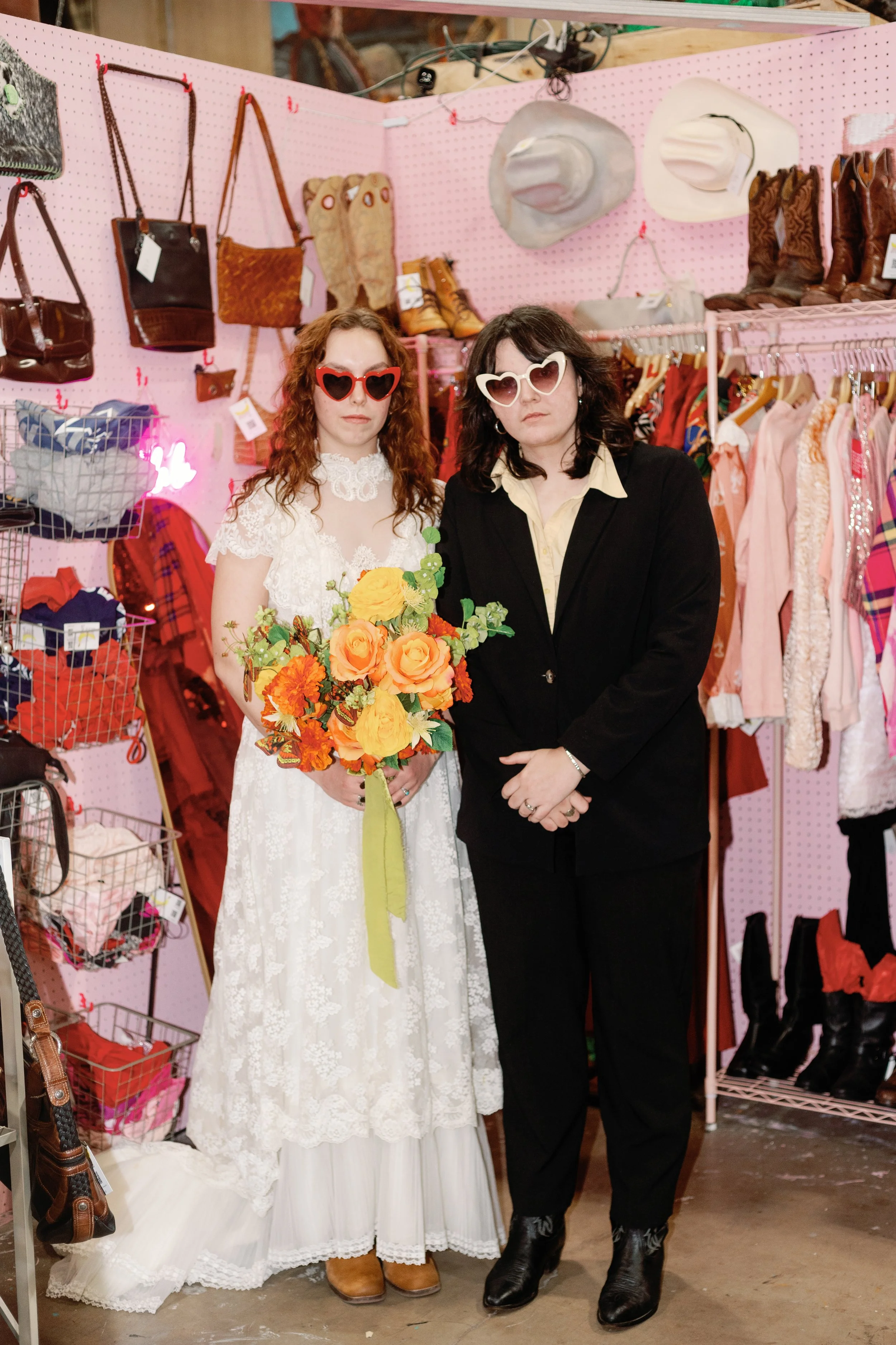 Two women wearing large heart-shaped sunglasses, standing in a vintage clothing store with pink pegboard walls and various accessories and boots on shelves behind them. One woman is in a white dress holding a bouquet of yellow and orange flowers, and