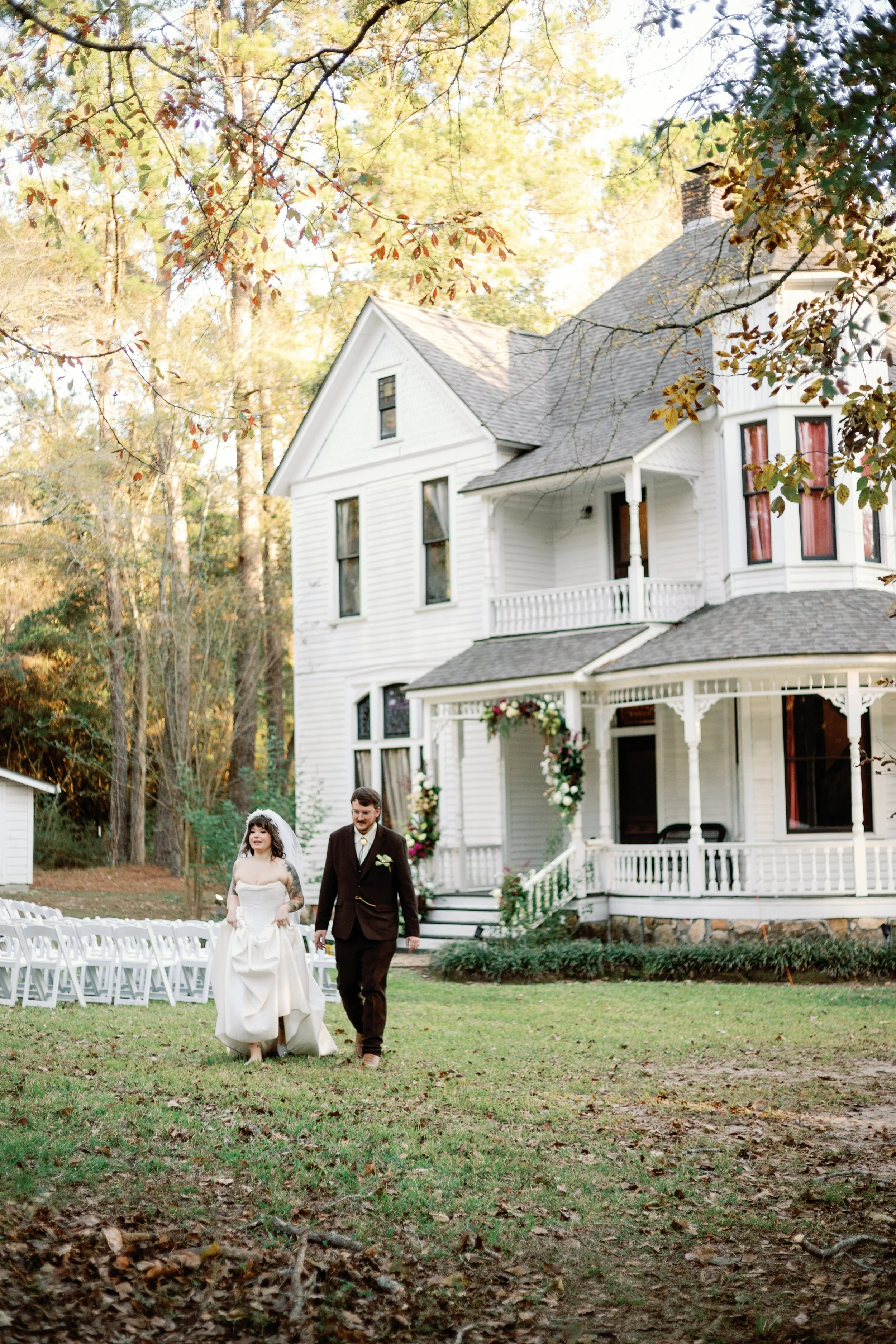 A couple walking together after their wedding during their reception outdoors in the fall with an old white two story house in the background.