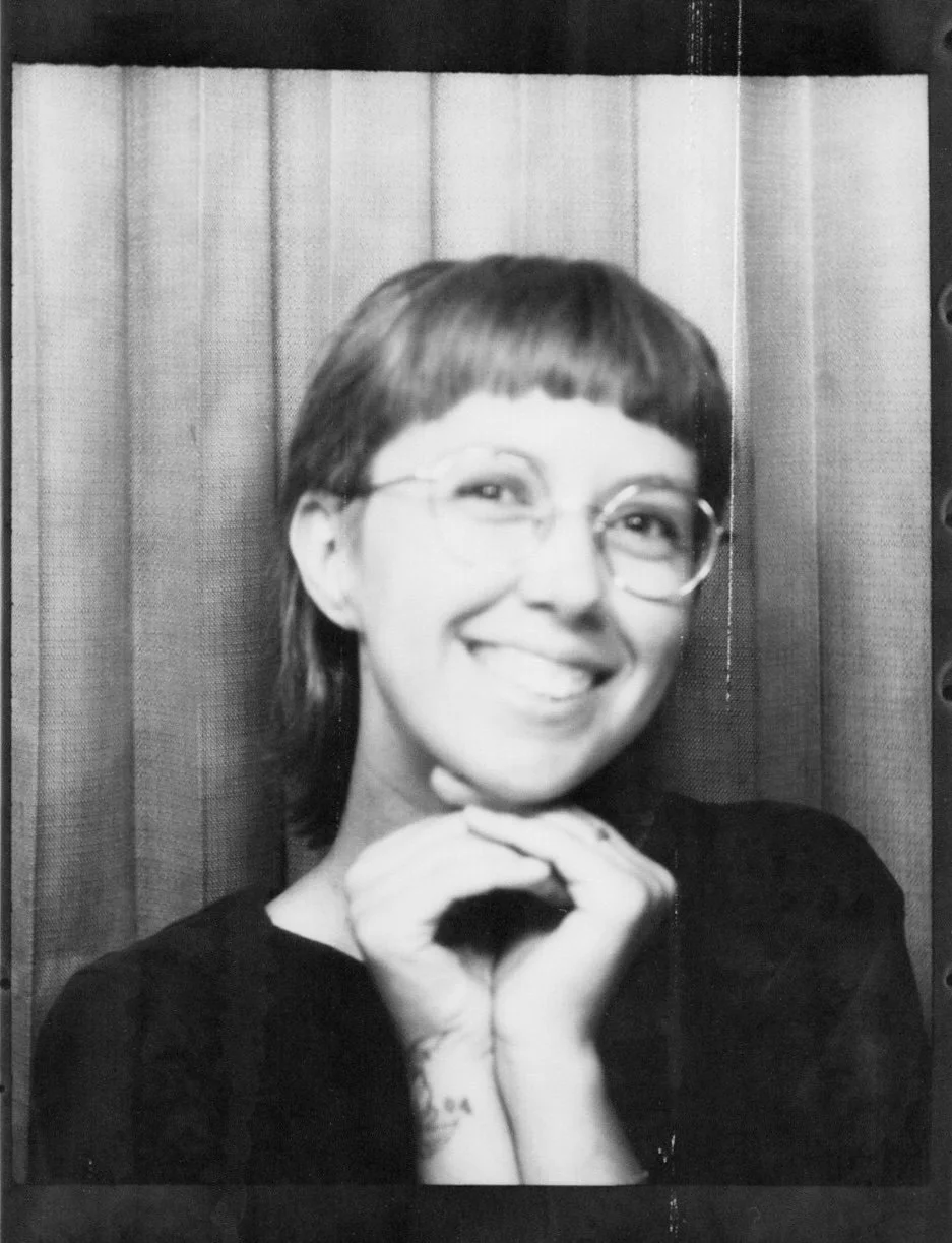 Black and white photo of a nonbinary person smiling with glasses, short hair, and a tattoo on their arm, posing in front of a wooden wall.