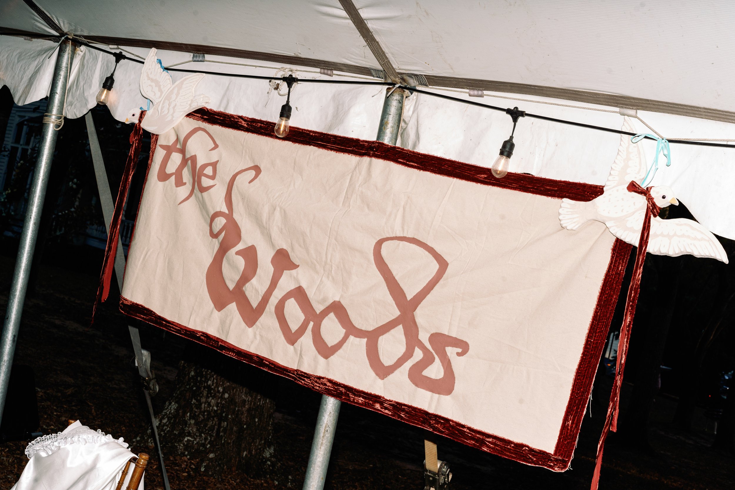 A flash photograph of a wedding sign that says "the woods" with custom carved dove banner holders during a wedding recpetion outdoors at night.