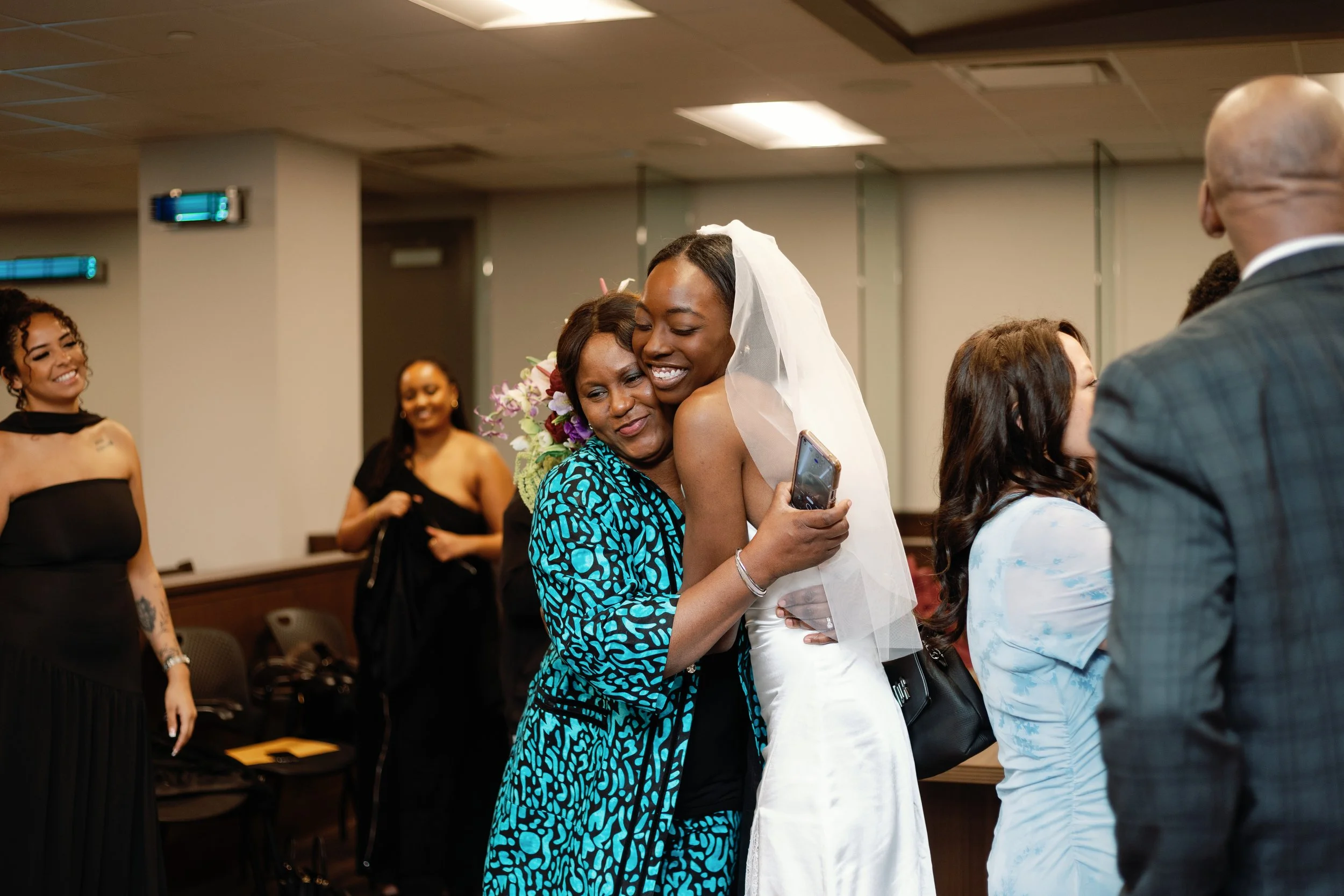 A woman in wedding attire is hugging another woman while holding a phone at her wedding reception, with other guests in the background.