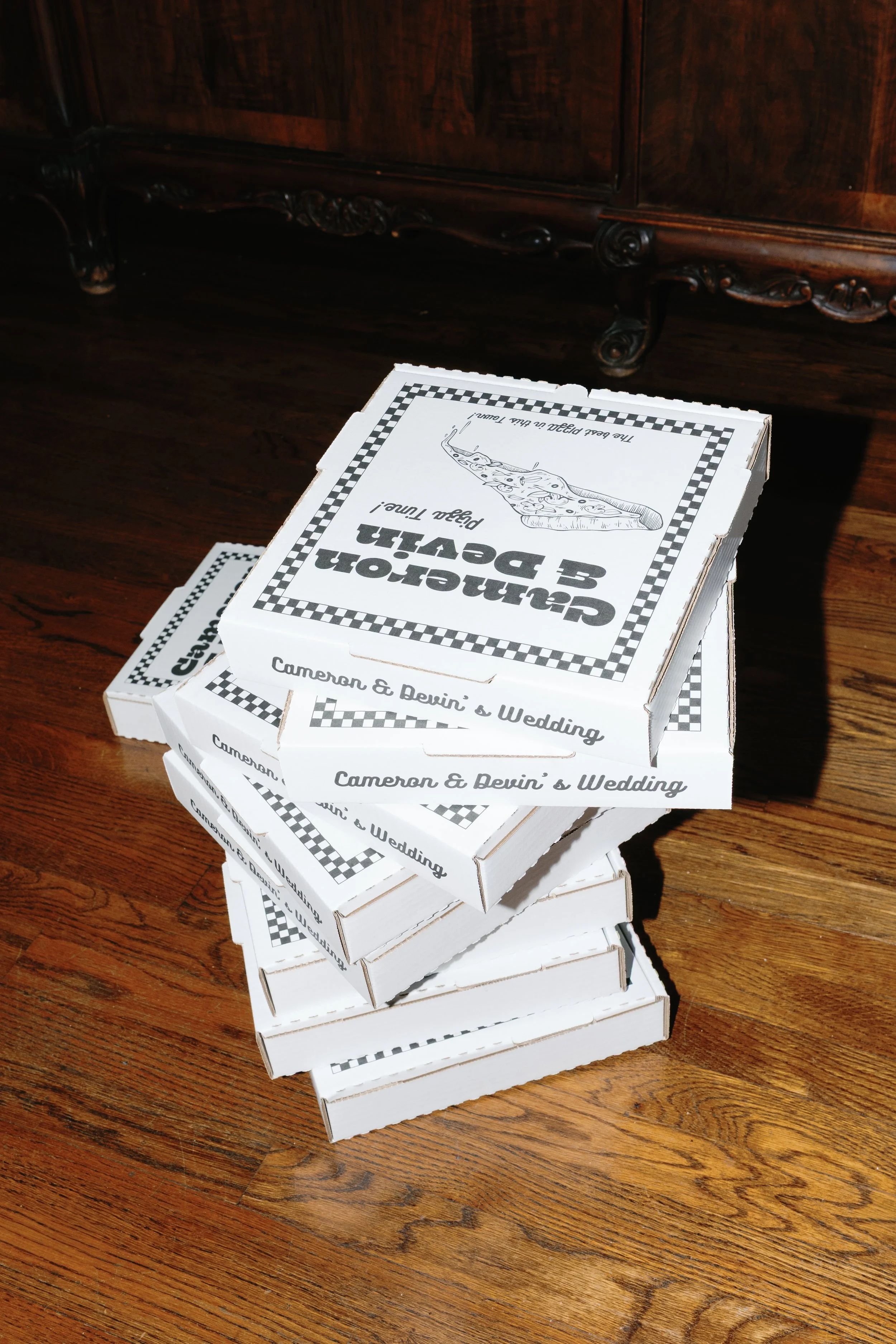 A stack of pizza boxes that are customized with a couples names on it sit rested on a warm toned wooden floor. The boxes will be used during their wedding reception.