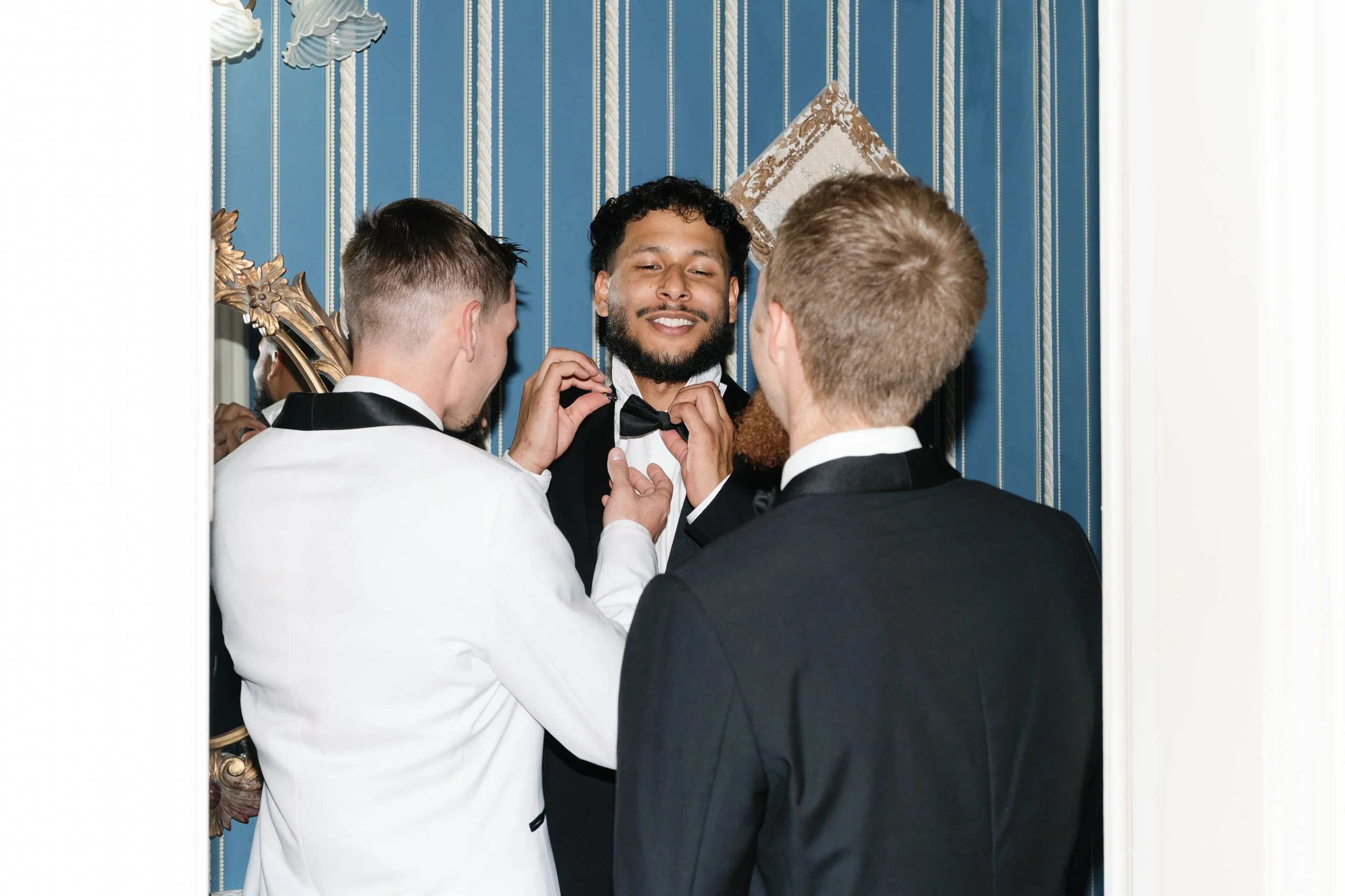 One of the Marriers adjusts the tie of a member of the wedding party with another member of the wedding party also in the bathroom.