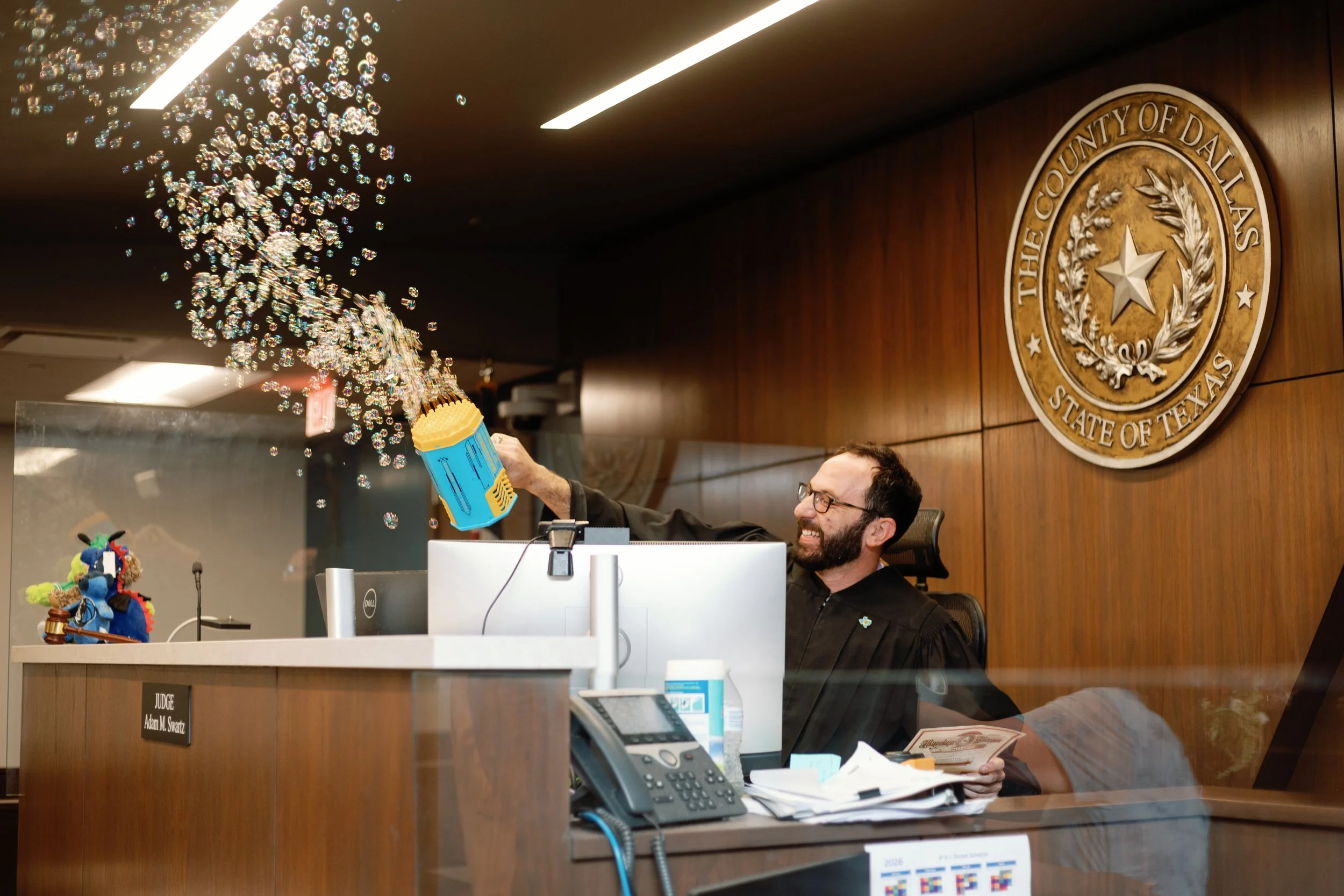 A judge sitting at a courtroom bench, smiling as he throws a party confetti cannon, with a large wooden seal of the County of Dallas, Texas, on the wall behind him.