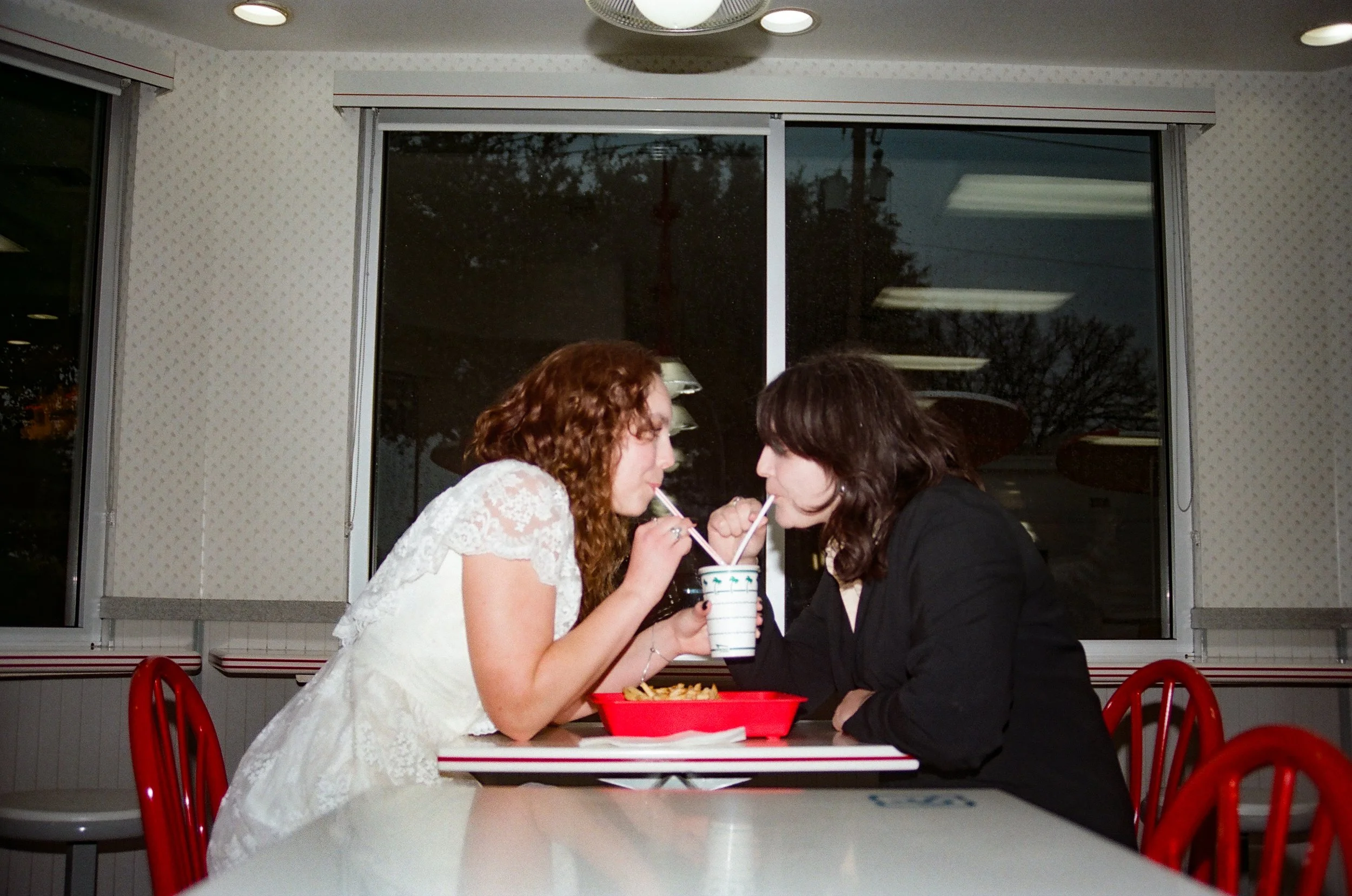 Two women sharing drinks from straws in a fast food restaurant, sitting across from each other at a table with a red tray of food. The background shows a large window with a view of trees and a dark sky.