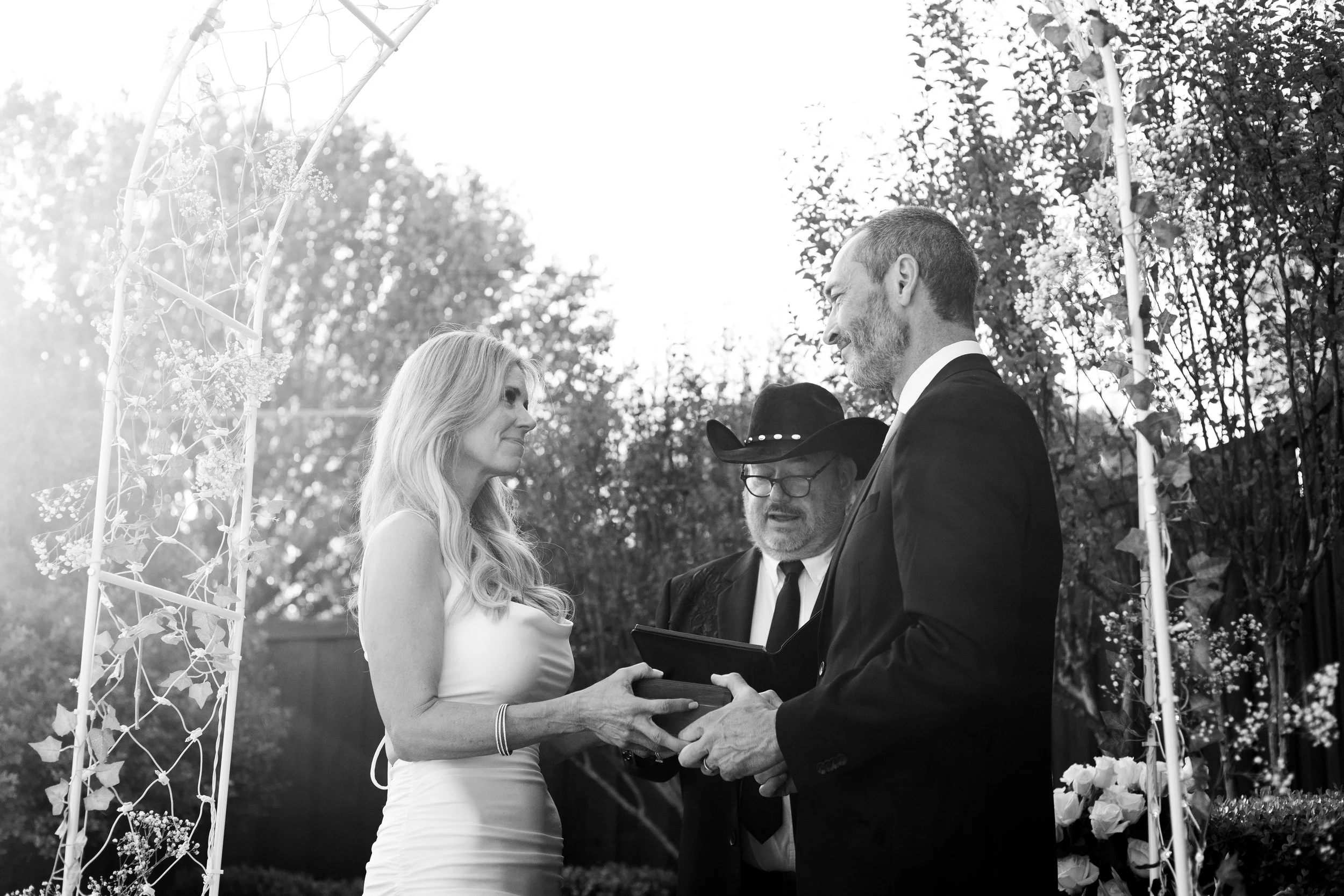 A woman and a man exchanging wedding vows during an outdoor wedding ceremony, officiated by a person wearing a black cowboy hat, with trees in the background.