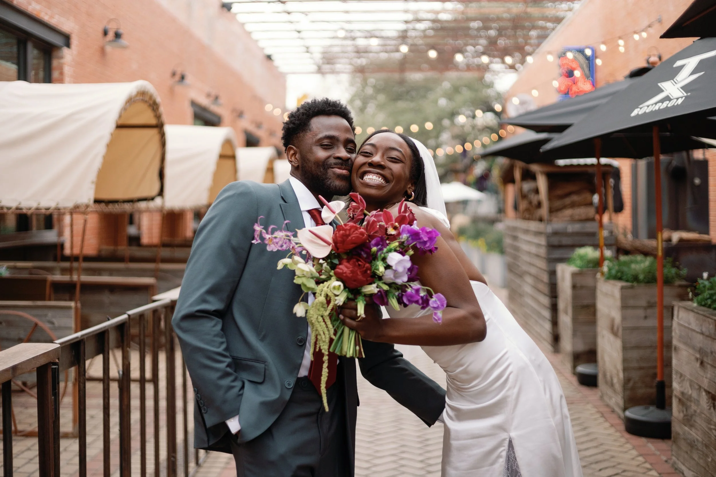 Happy couple embracing and smiling, with a woman holding a colorful bouquet, outdoors restaurant patio with string lights, umbrellas, and planters.