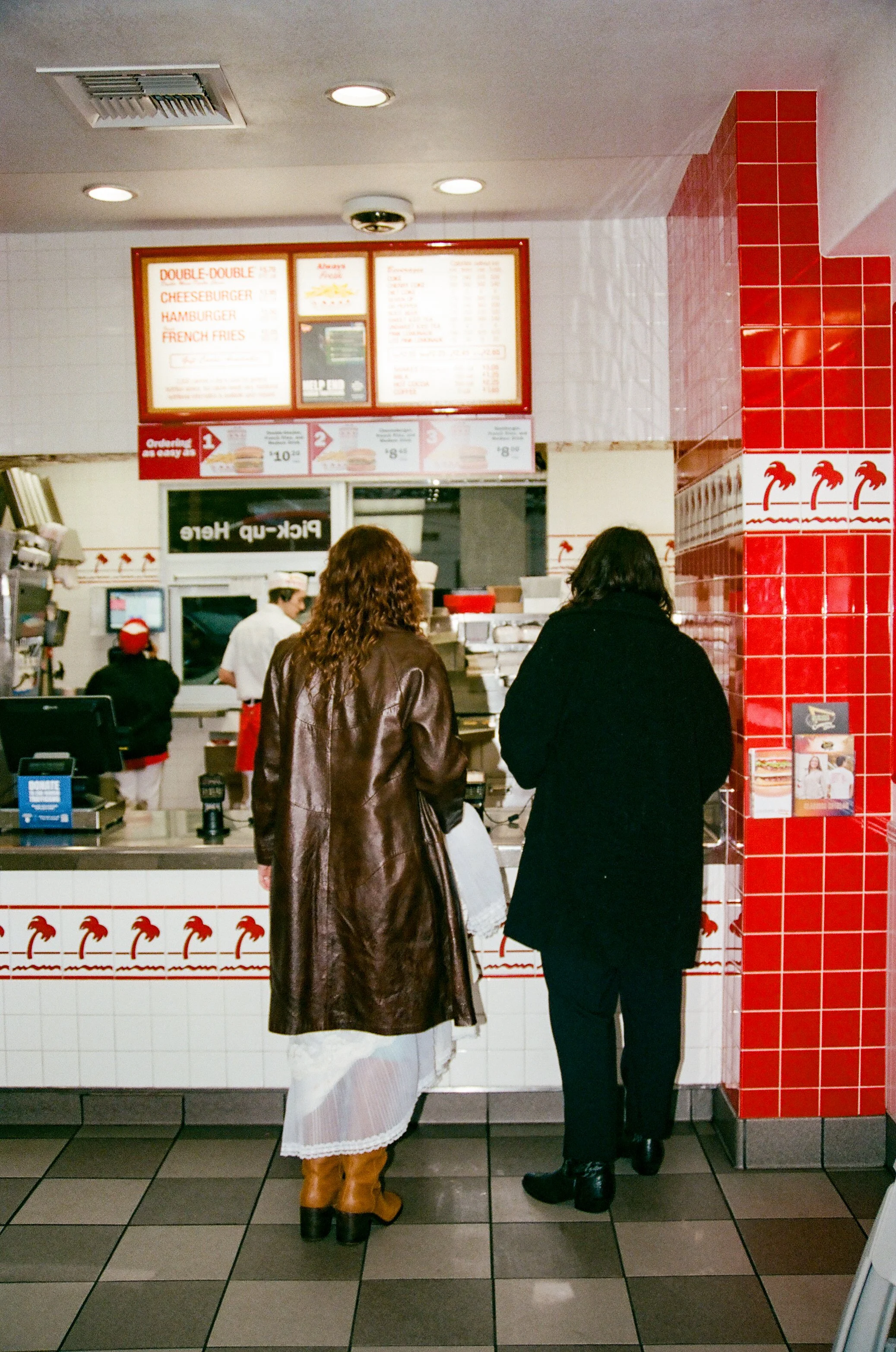 Two women standing at the counter of a fast food restaurant, ordering food, with employees visible inside the kitchen.