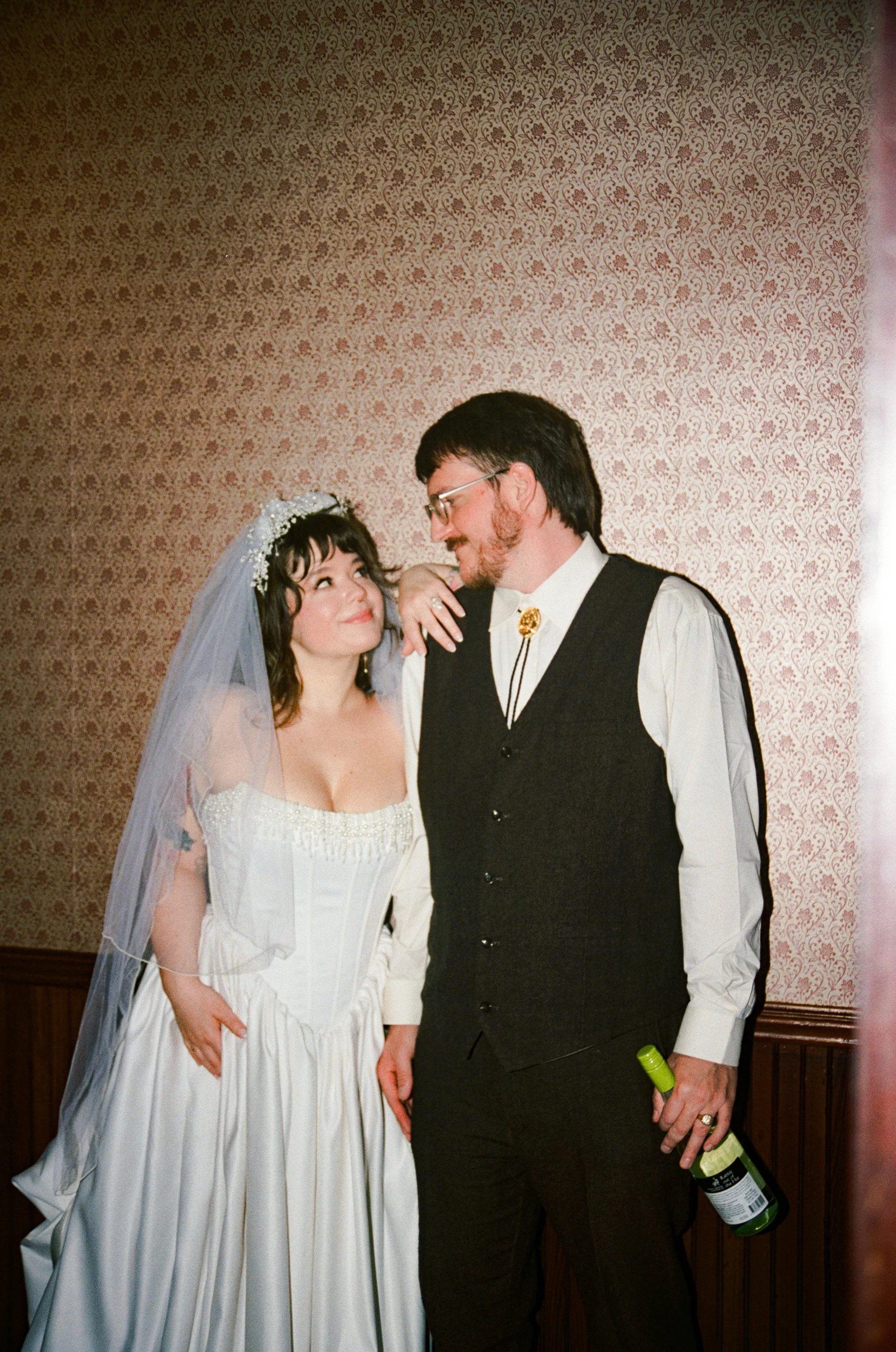 A newlywed couple lean on eachother and stare into eachothers eyes while smiling in a hallway with patterned vintage wallpaper on the wall.