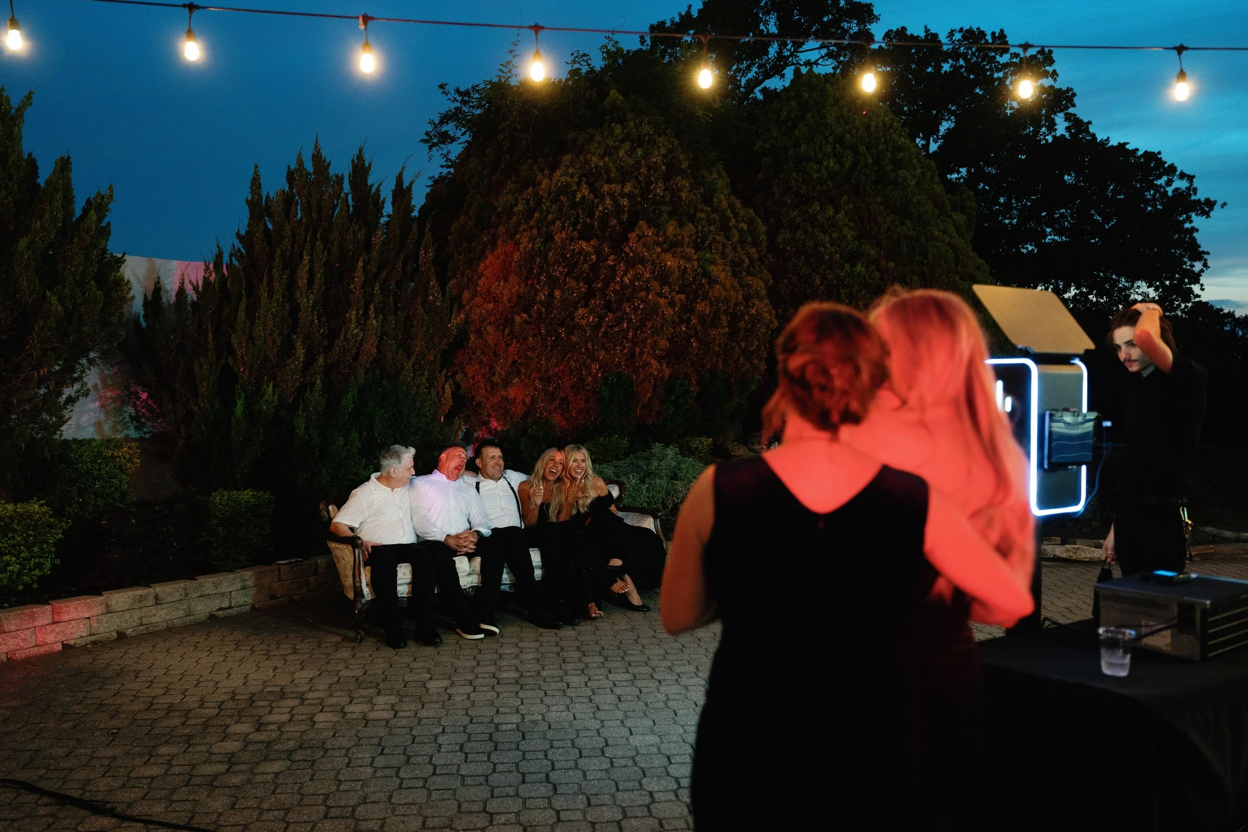 A marrier and their relative watch a group of family members pose while taking pictures at a photobooth during their outdoor wedding reception at night.