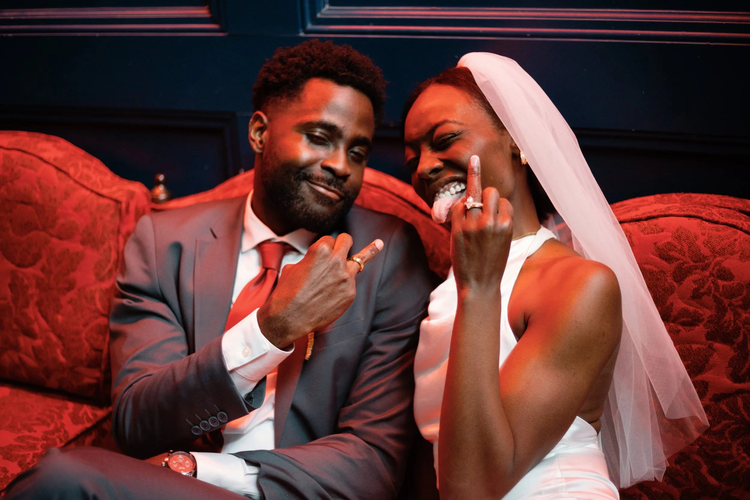 A newlywed couple sitting on a red velvet couch, dressed in wedding attire, making playful gestures and smiling.