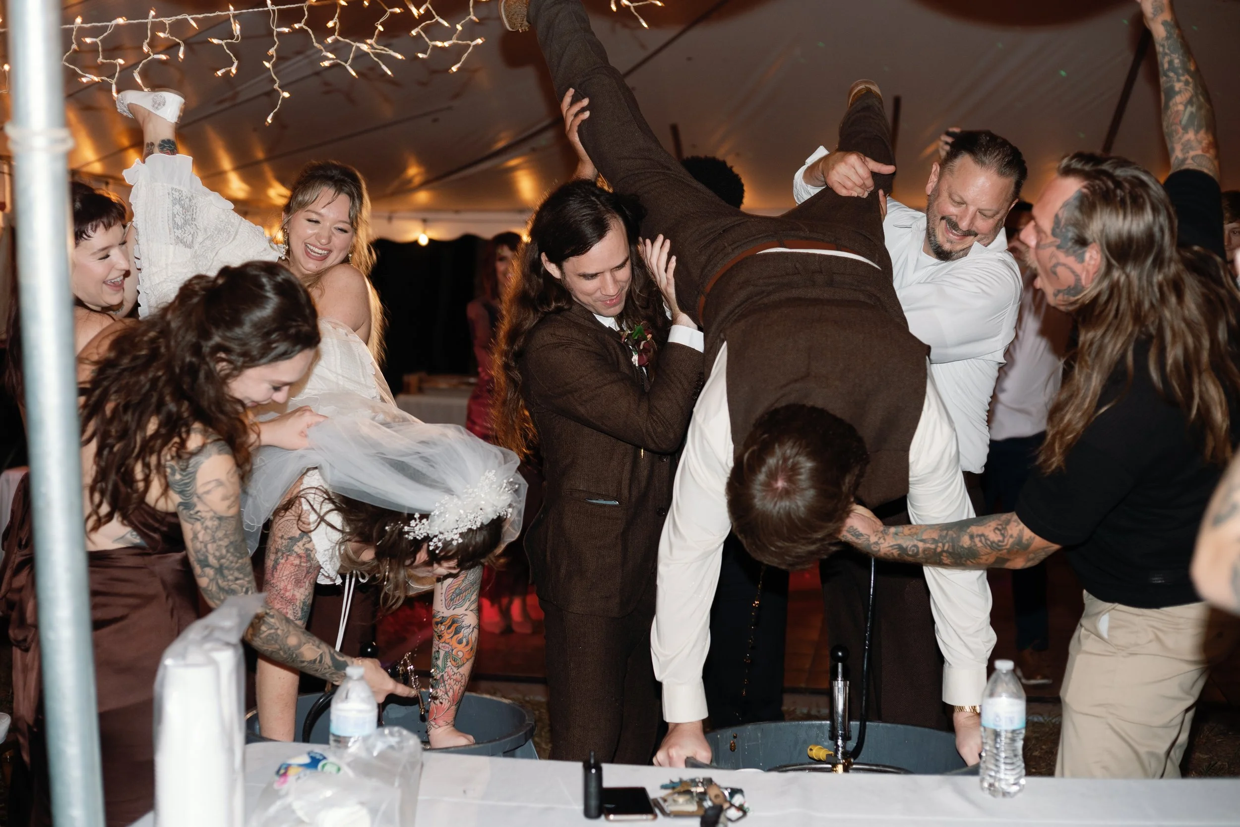 Newlyweds are being held in the air by their wedding party while they do a keg stand together at their outdoor wedding recpetion at night.