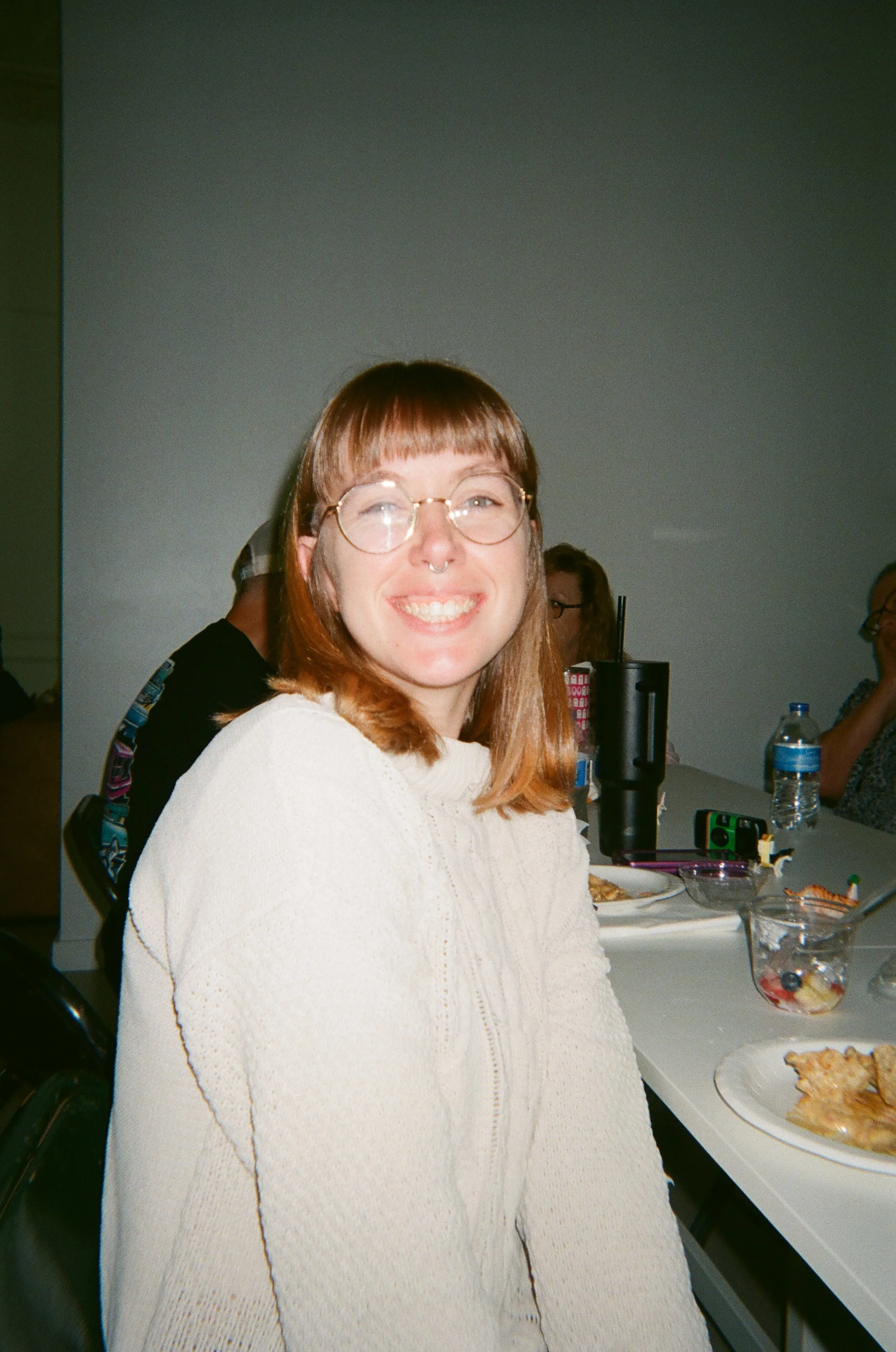 Smiling woman with glasses and light brown hair at a gathering, sitting at a table with plates of food, drinks, and other people in the background.
