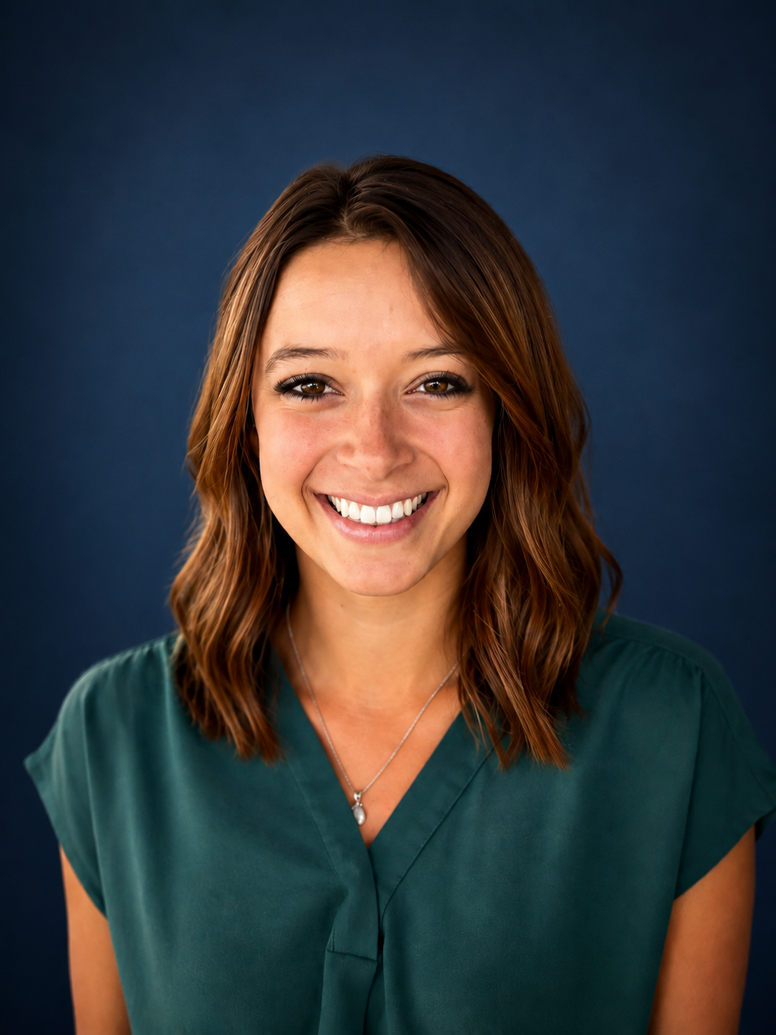 Portrait of a smiling woman with shoulder-length brown hair, wearing a teal top, standing in front of a wooden background.
