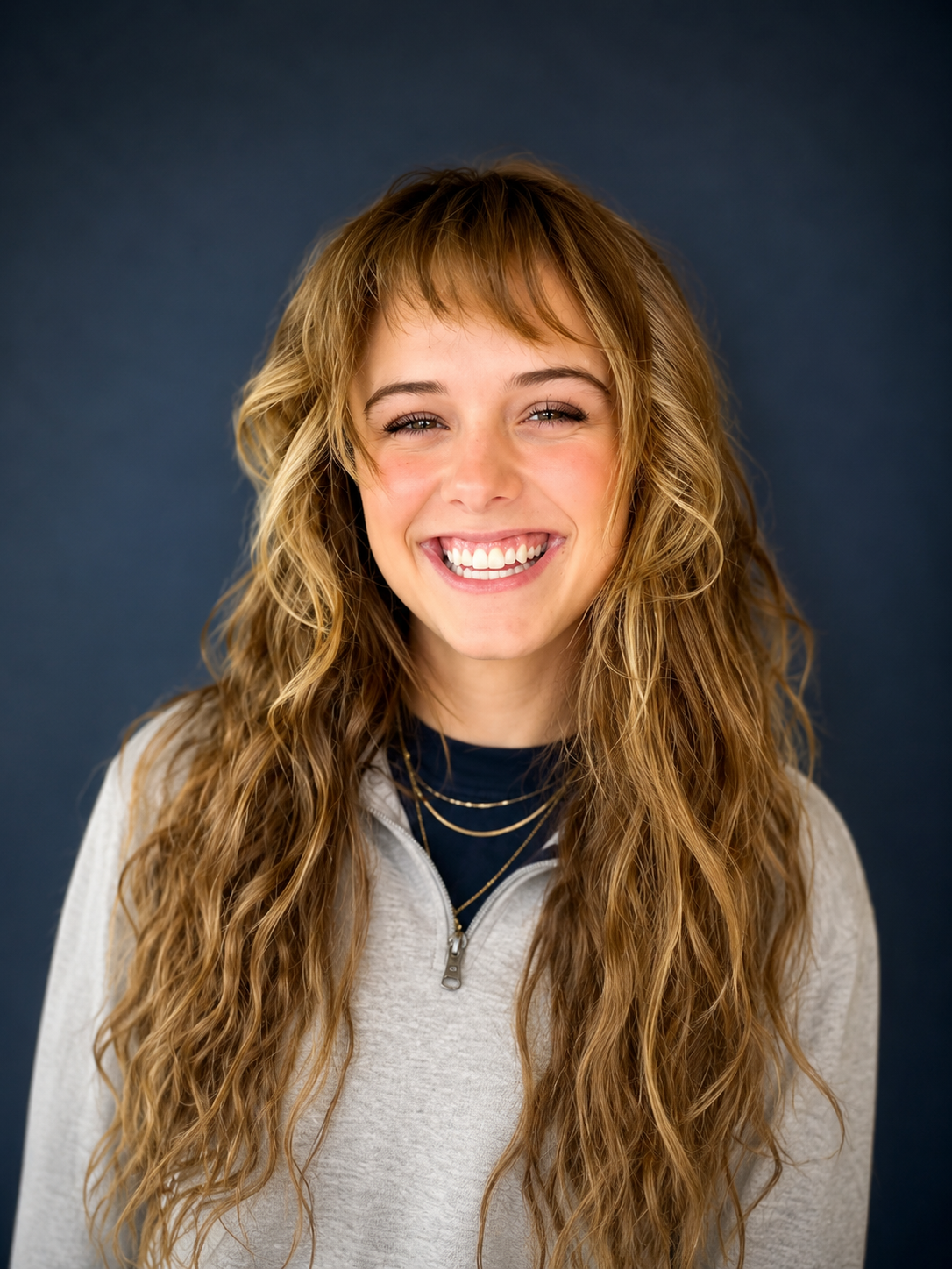 A young woman with long, wavy, light brown hair and bangs, smiling with her teeth showing, standing in front of a pinkish-brown wooden wall.