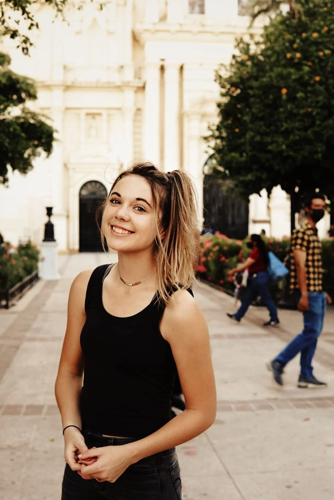 A young woman with shoulder-length hair smiling outdoors on a city street, wearing a black tank top and a small necklace, with other pedestrians and greenery in the background.
