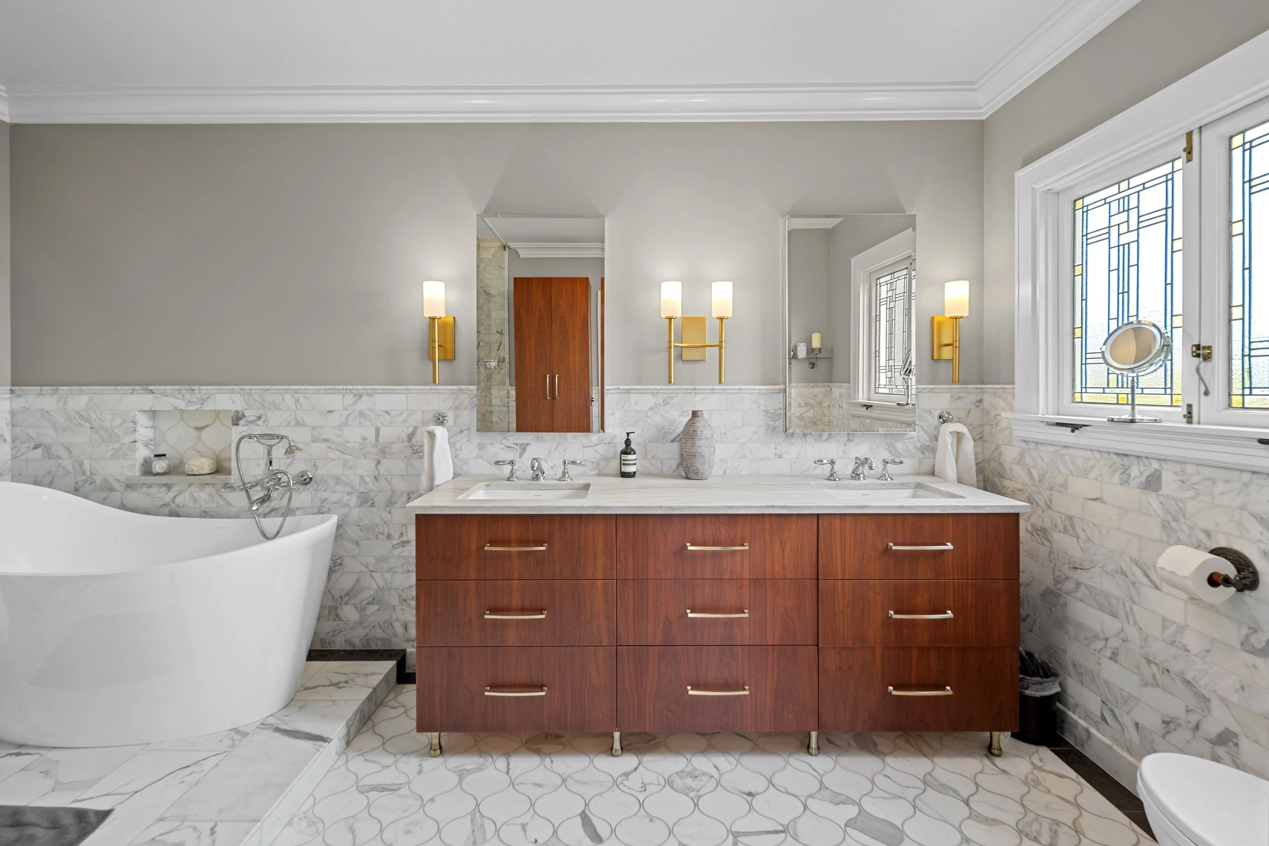 Bathroom with a white bathtub, a wooden vanity with two sinks, marble countertops, and marble wall tiles, illuminated by wall-mounted yellow light fixtures. Features large windows with decorative stained glass, a mirror, and bathroom accessories.